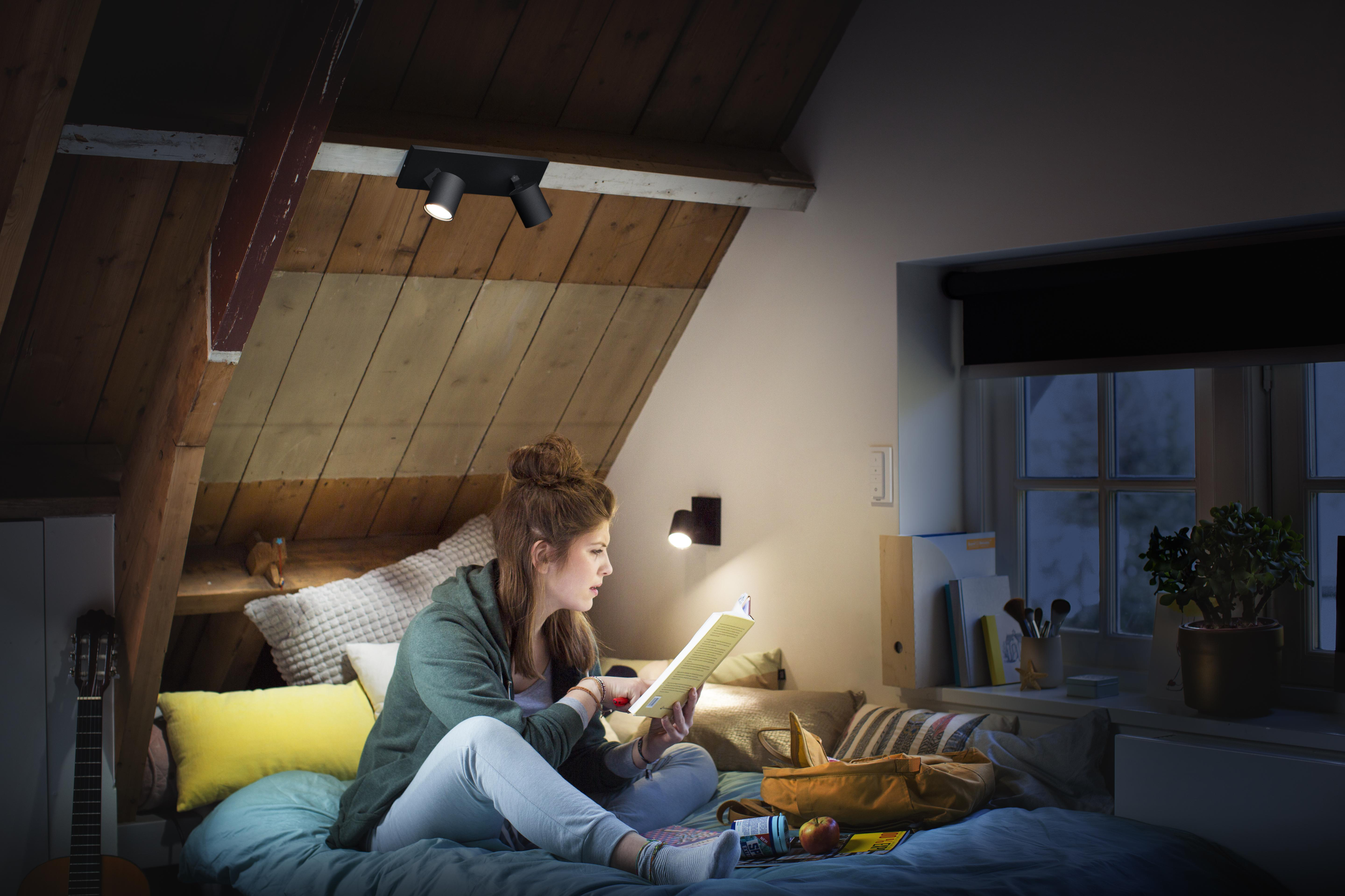 Frau liest in einem Zimmer, das von Lampen beleuchtet wird. Es gibt ein Fenster, ein Bett und eine Gitarre in der Nähe.