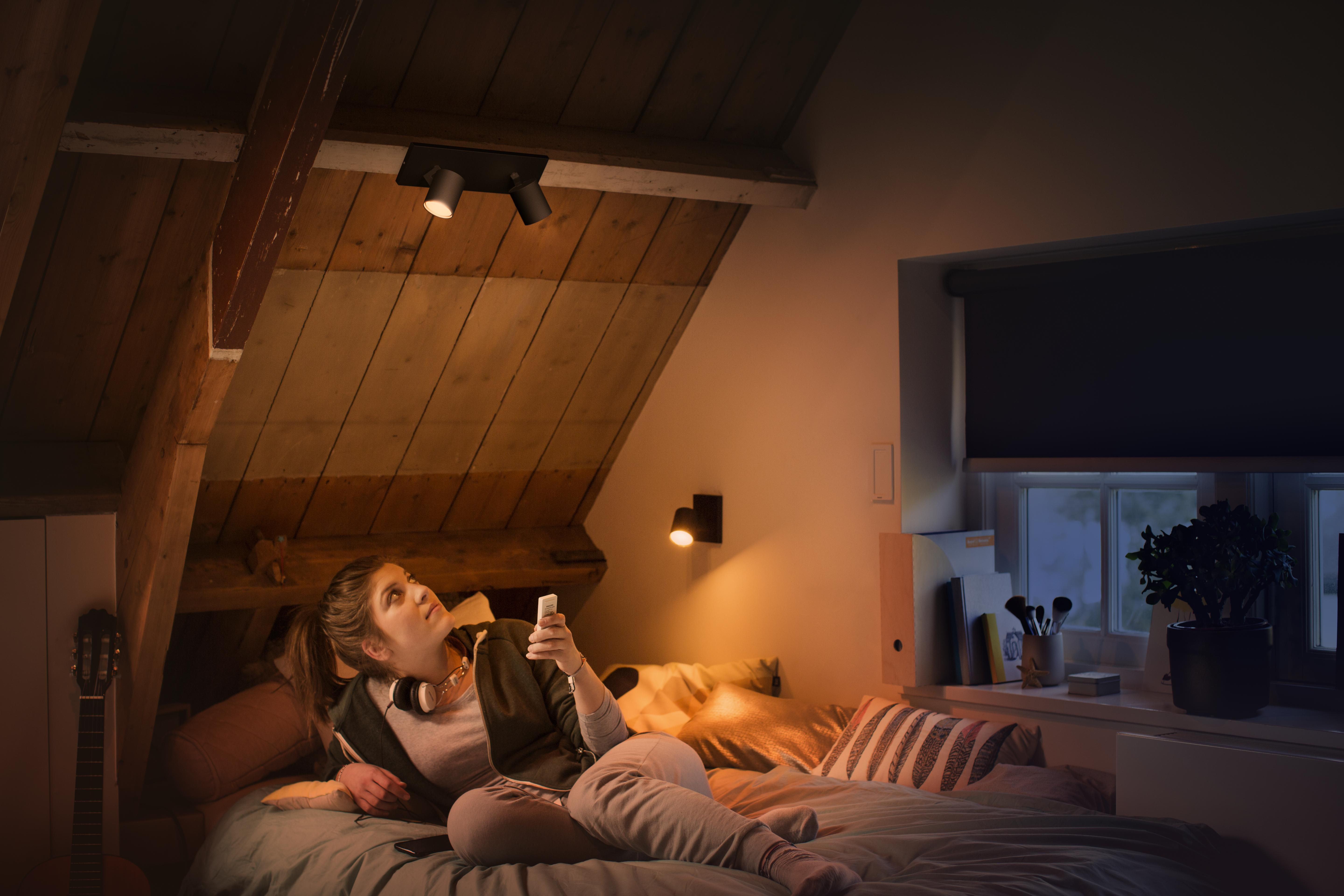 Eine Frau in einem Schlafzimmer blickt auf eine Deckenleuchte und hält eine Fernbedienung.