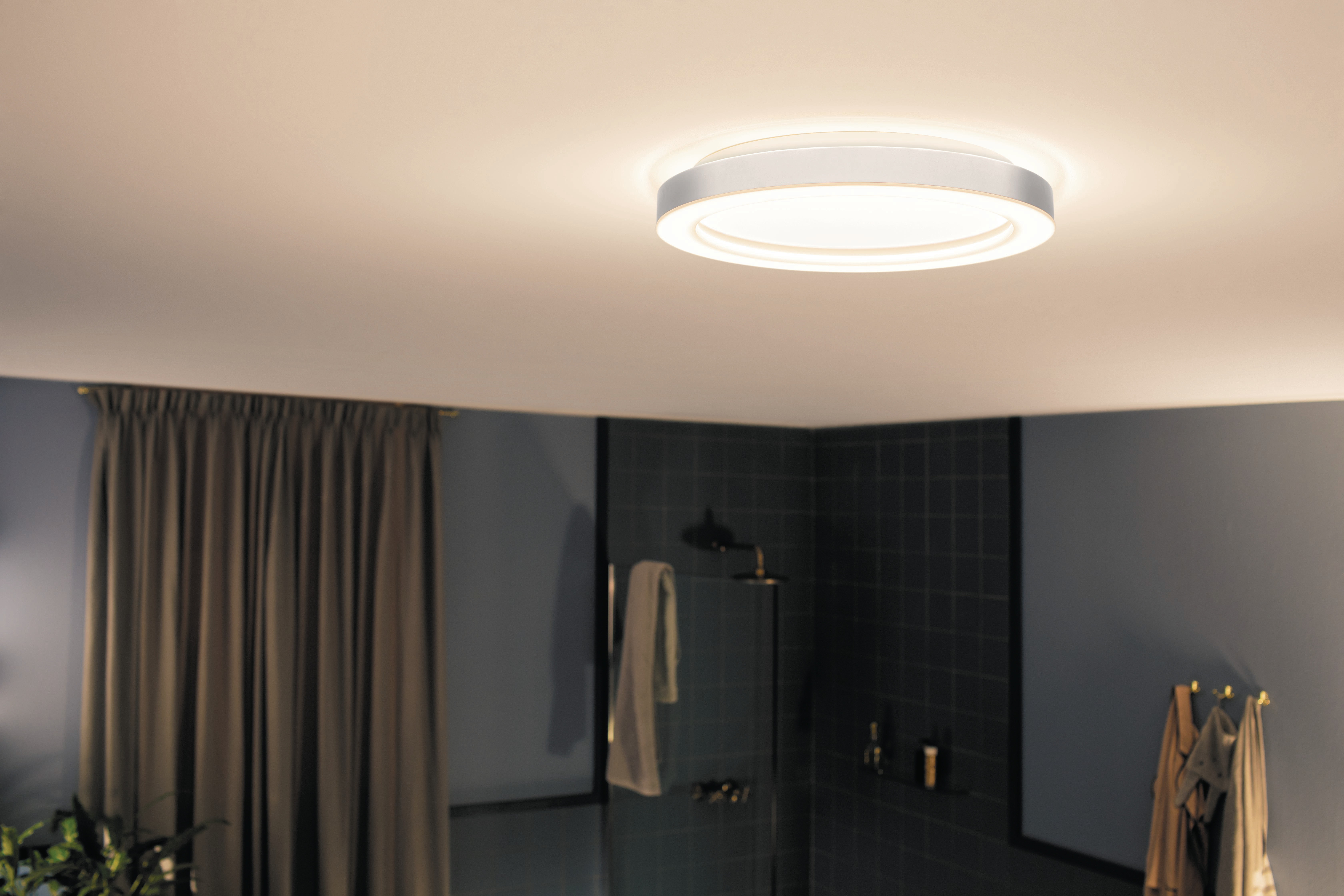 Eine Badezimmerdecke mit einer runden, beleuchteten Lampe. Dunkle Vorhänge links.