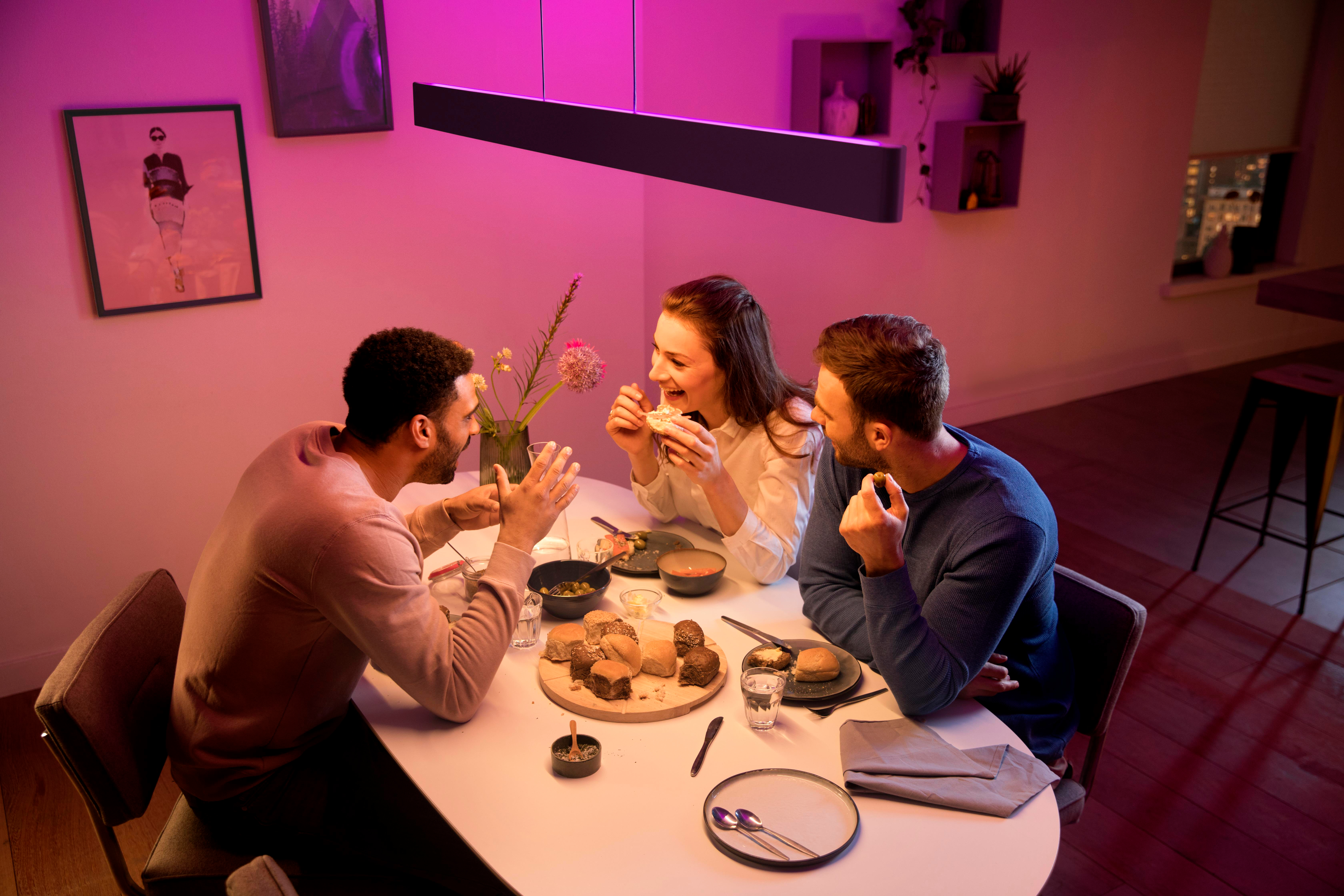 Drei Personen lachen und essen an einem Tisch unter lila Umgebungslicht. Speisen und Getränke vorhanden.