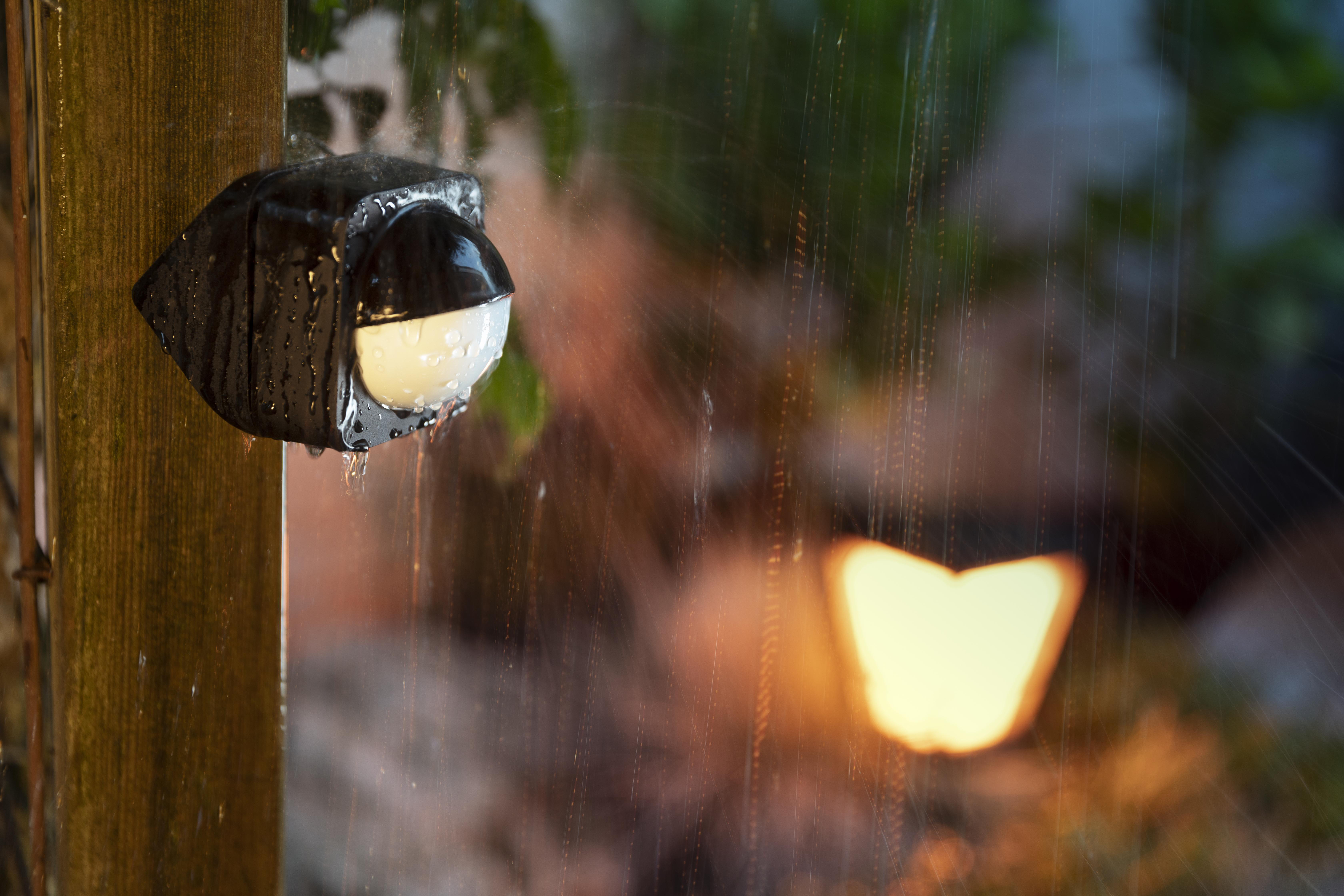 Ein schwarzer Außenlichtsensor an einem Holzpfosten mit Regen und Licht im Hintergrund.