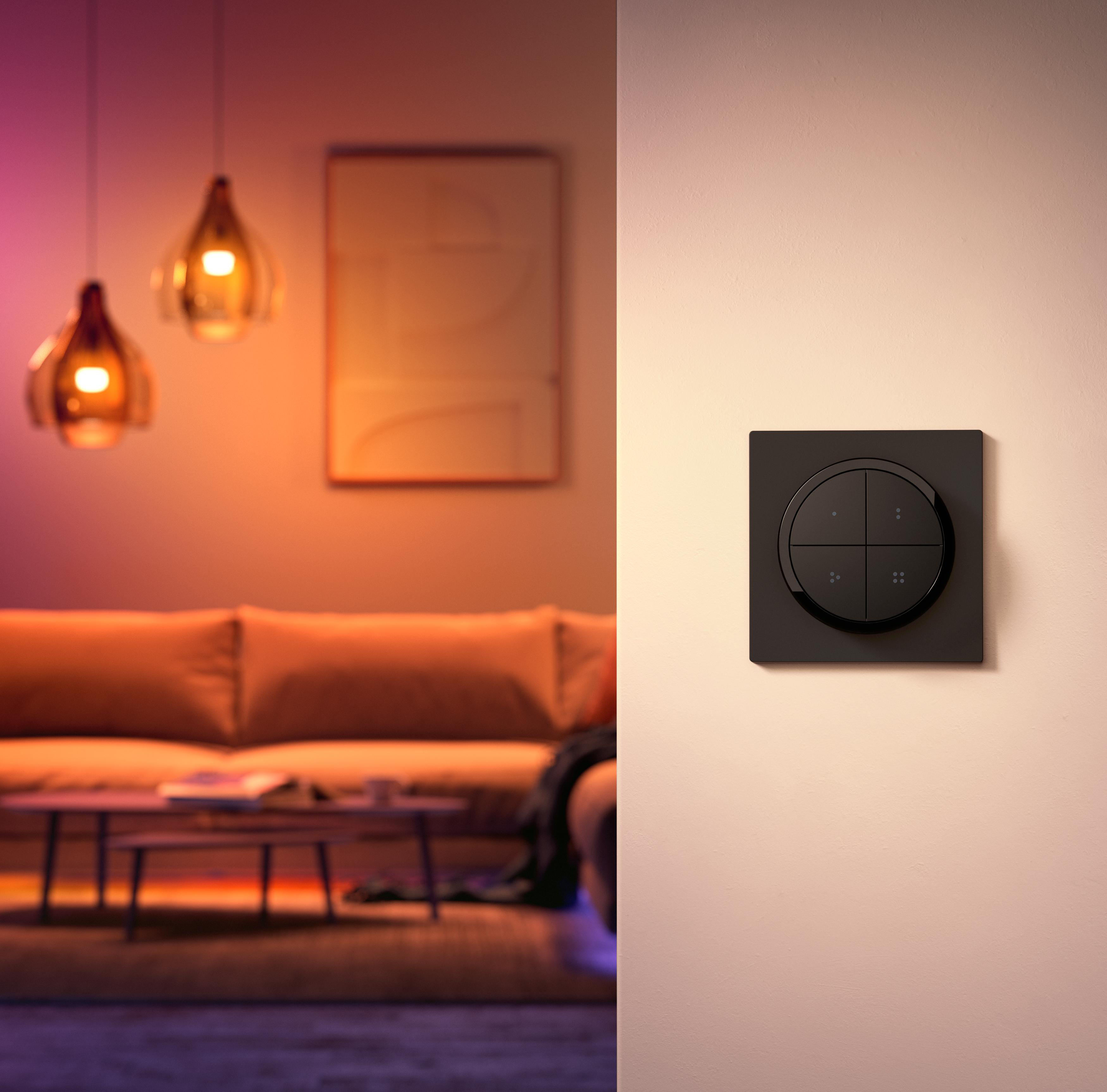 Ein an der Wand montierter schwarzer Smart-Lichtschalter mit vier Tasten. Im Hintergrund ein Raum mit Lichtern.