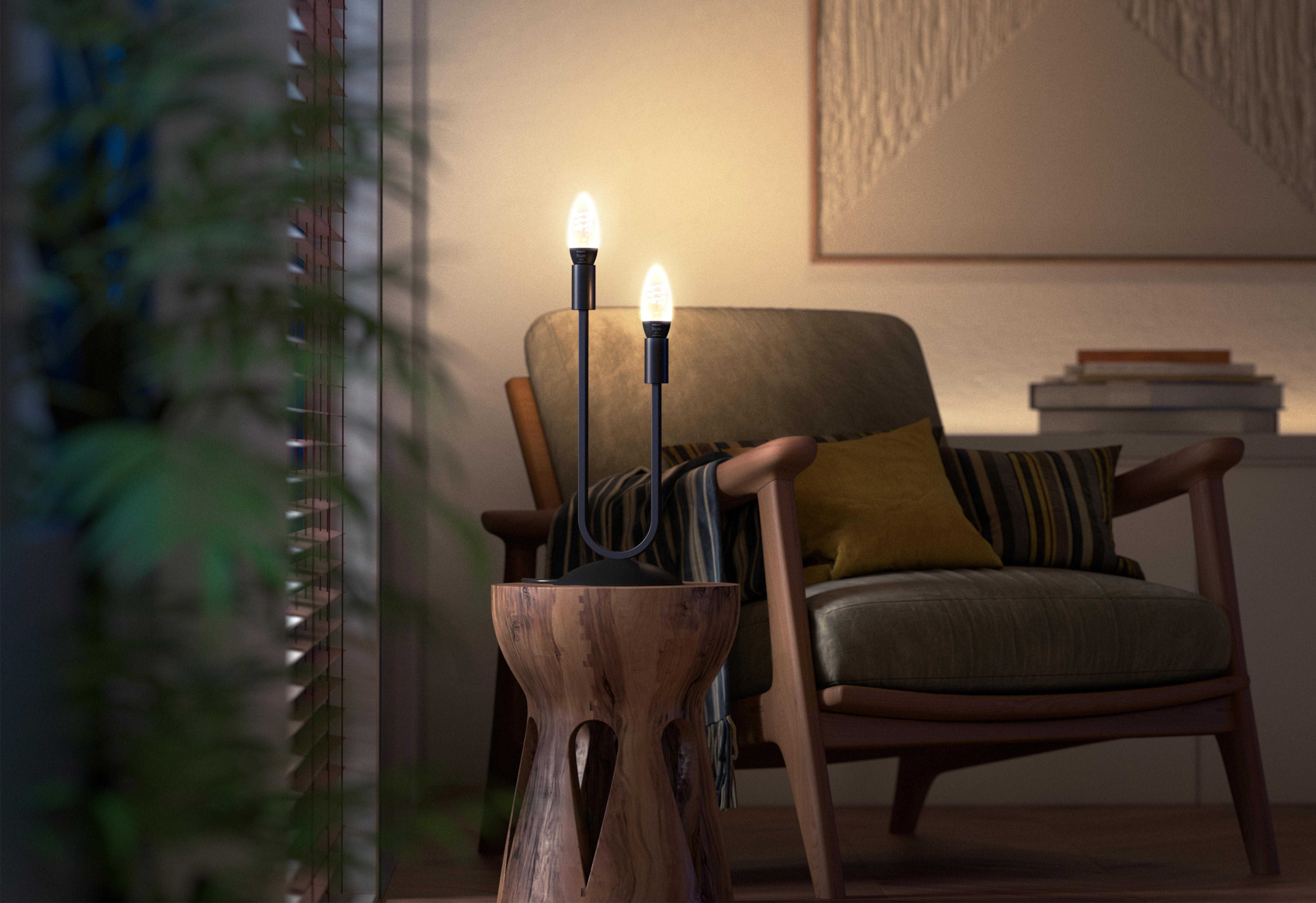 Eine Lampe mit zwei leuchtenden Glühbirnen steht auf einem Holzschemel. Im Hintergrund ein Sessel mit Kissen.