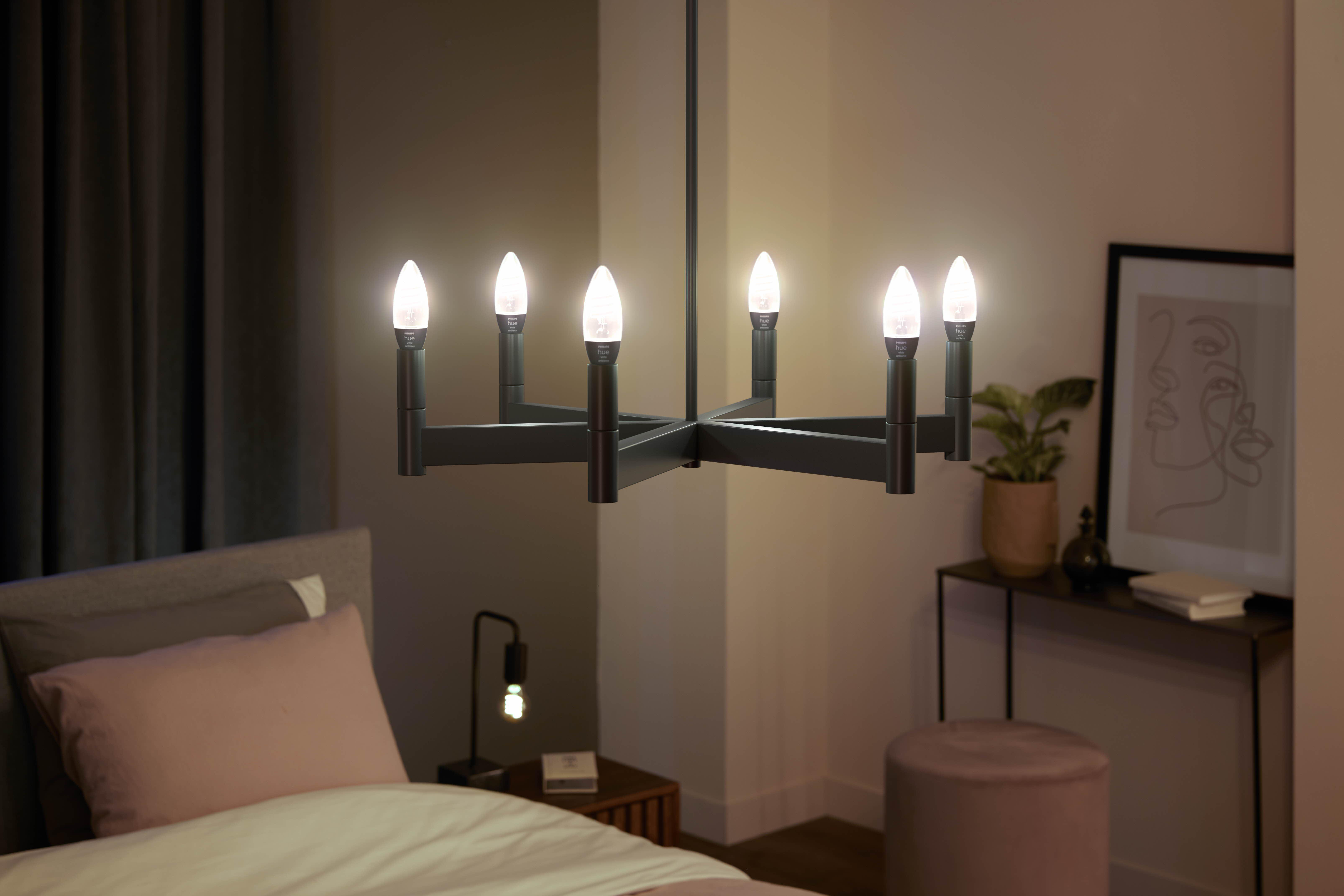 Ein Kronleuchter mit sieben brennenden Glühbirnen hängt über einem Bett mit rosa Kissen. Eine Lampe und ein Beistelltisch sind ebenfalls sichtbar.