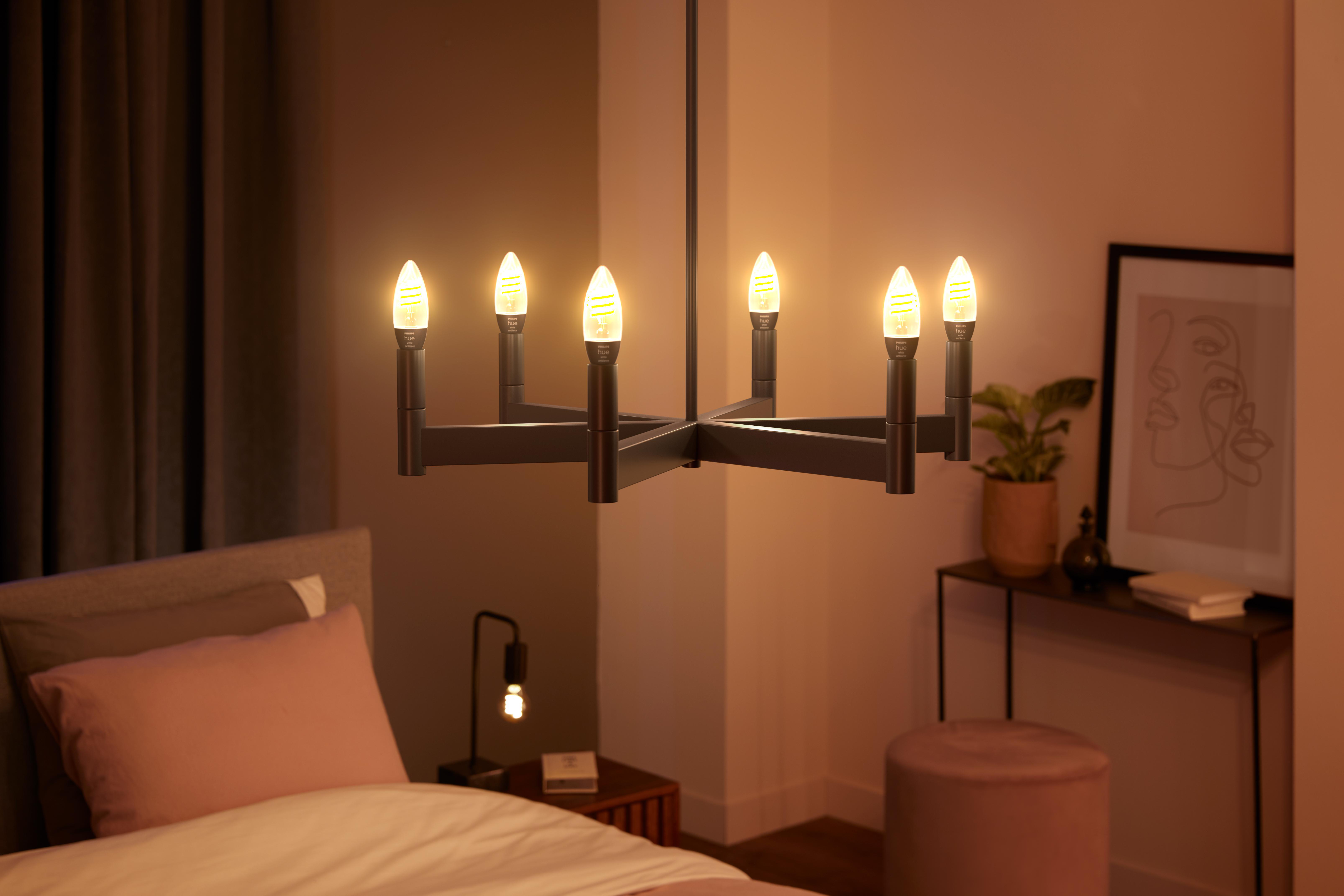 Ein Kronleuchter mit sechs leuchtenden Glühbirnen hängt über einem Bett. Eine Lampe und Kunstwerke stehen auf dem Beistelltisch.