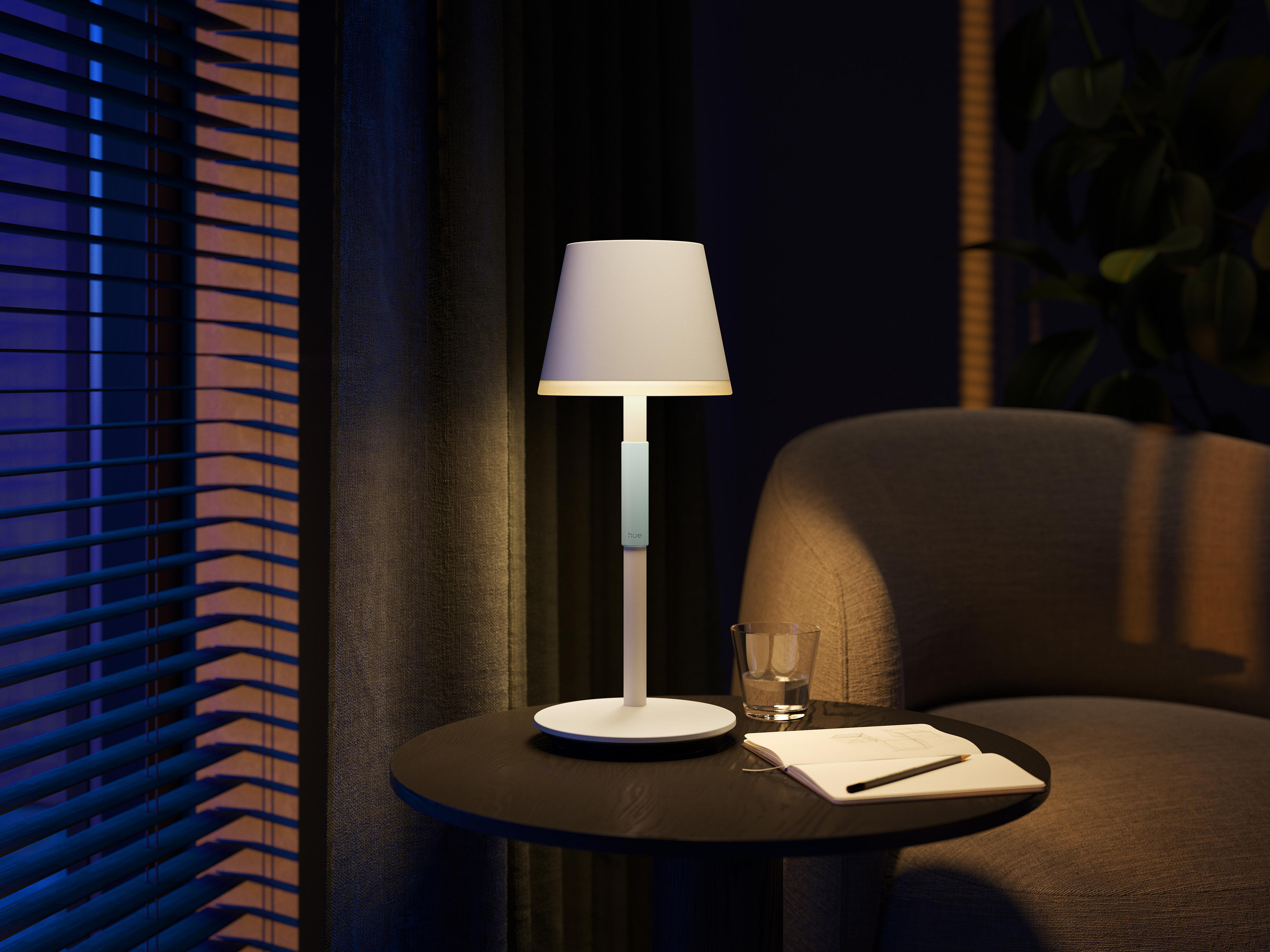 Eine beleuchtete Tischlampe auf einem Tisch mit einem Buch und einem Glas Wasser. Ein Stuhl steht daneben.