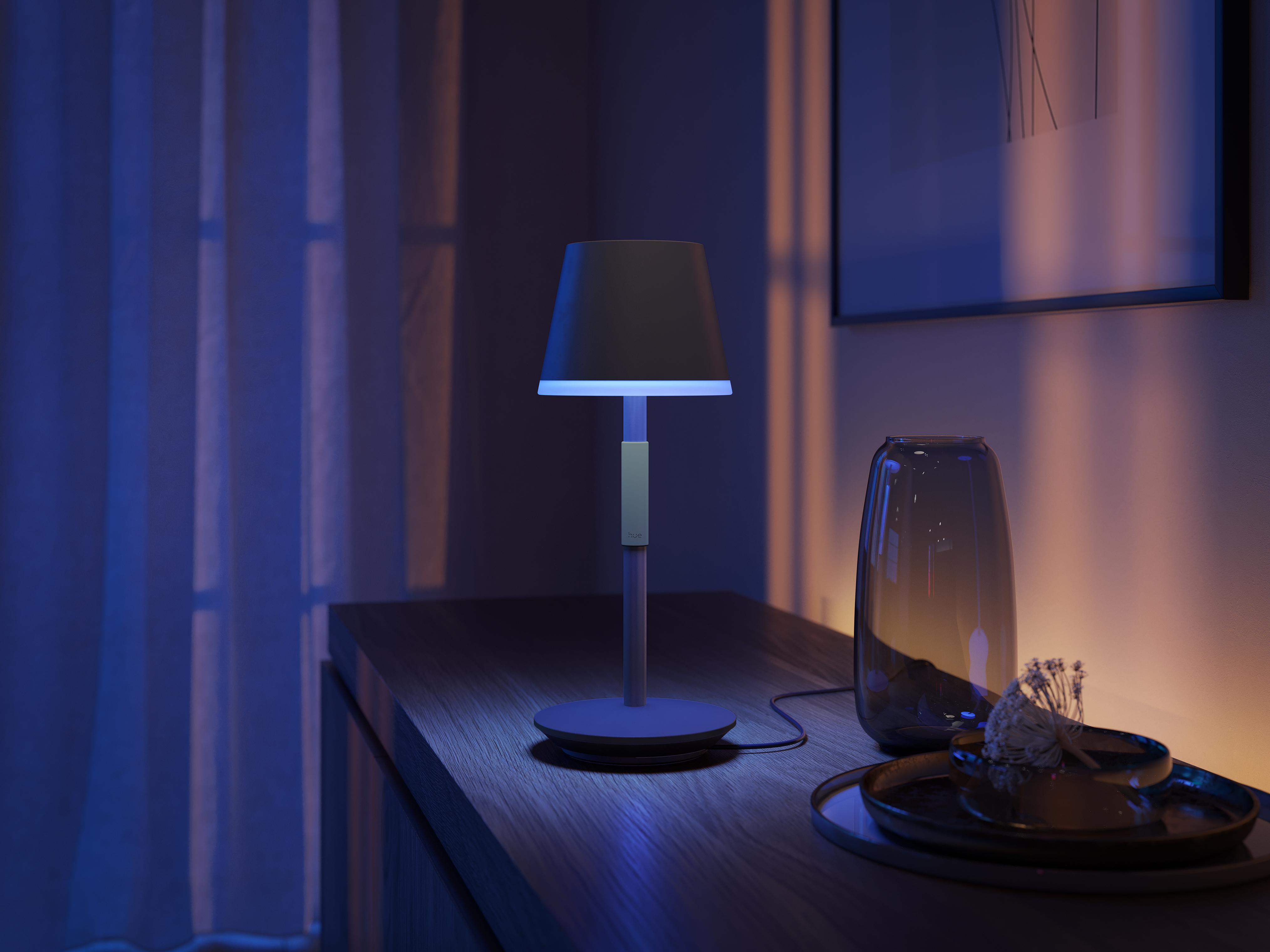 Eine Lampe auf einem Tisch. Eine Vase und ein Tablett daneben. Der Raum ist blau beleuchtet.