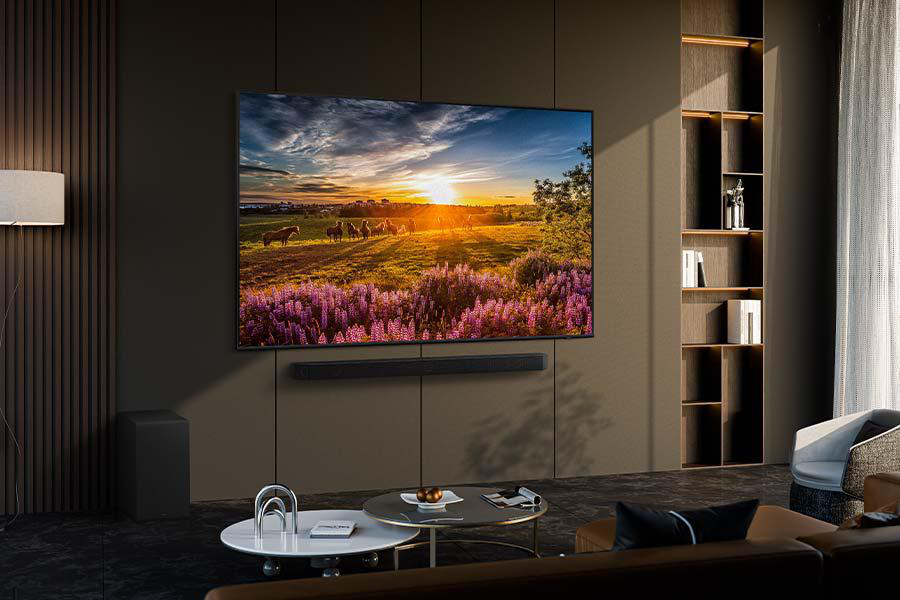 Ein großer Fernseher zeigt eine Sonnenuntergangsszene. Eine Soundbar befindet sich darunter, ein Subwoofer steht am Boden.