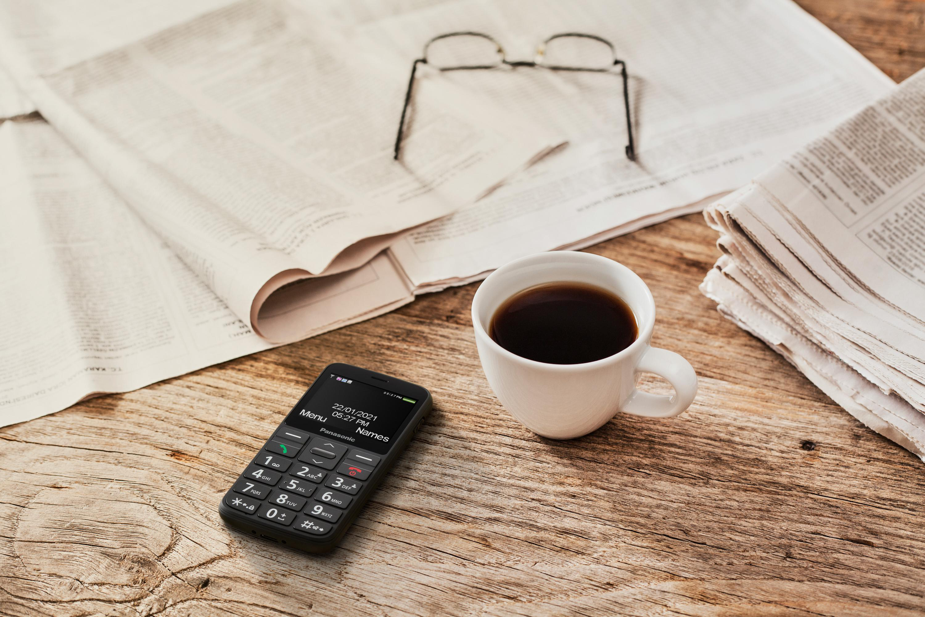 Ein schwarzes Mobiltelefon mit Anzeige, Kaffeetasse, Brille, Zeitungen auf einem Holztisch.