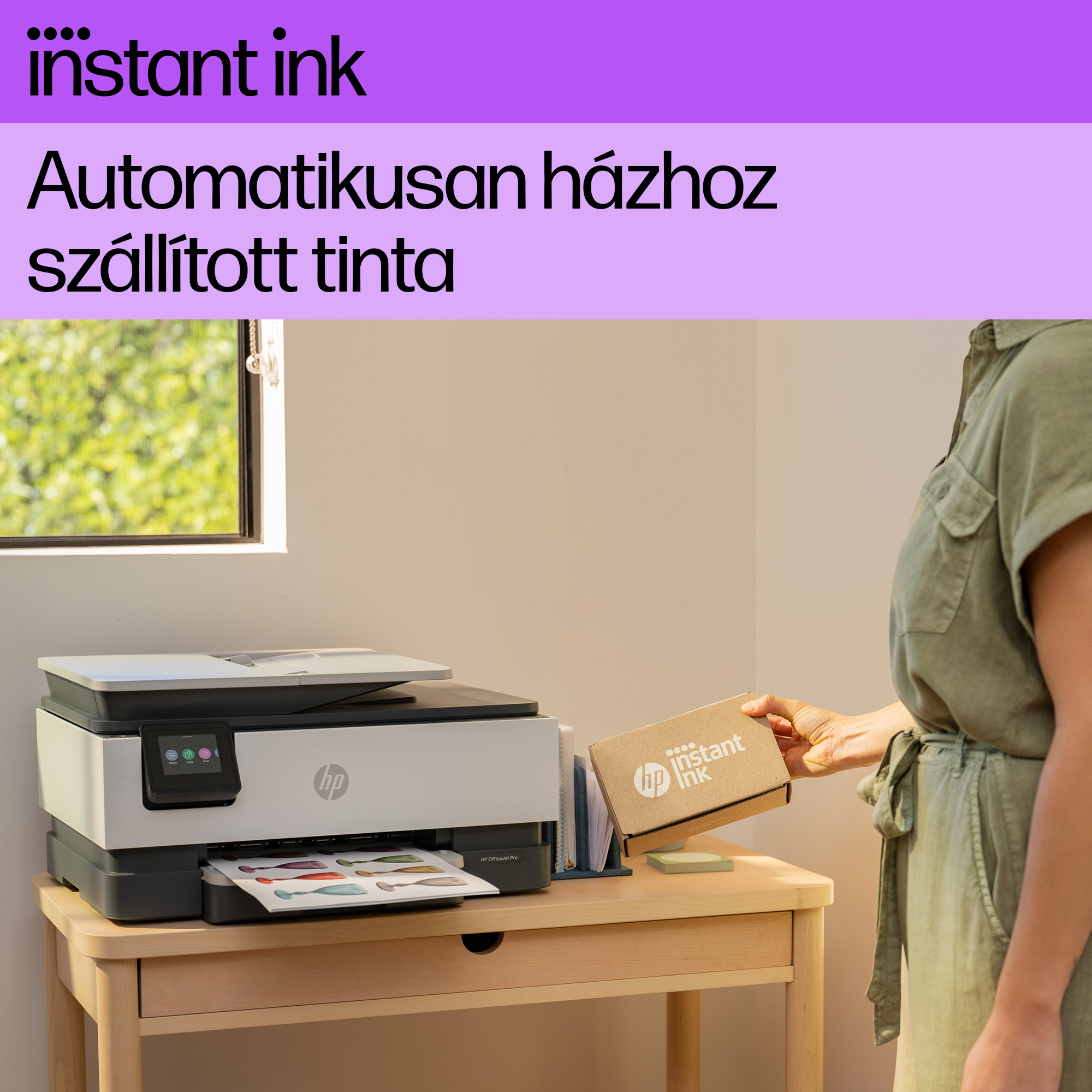 Egy nő egy 'Instant Ink' feliratú dobozt kap egy nyomtató mellett, egy fa asztalon, az ablak mellett.