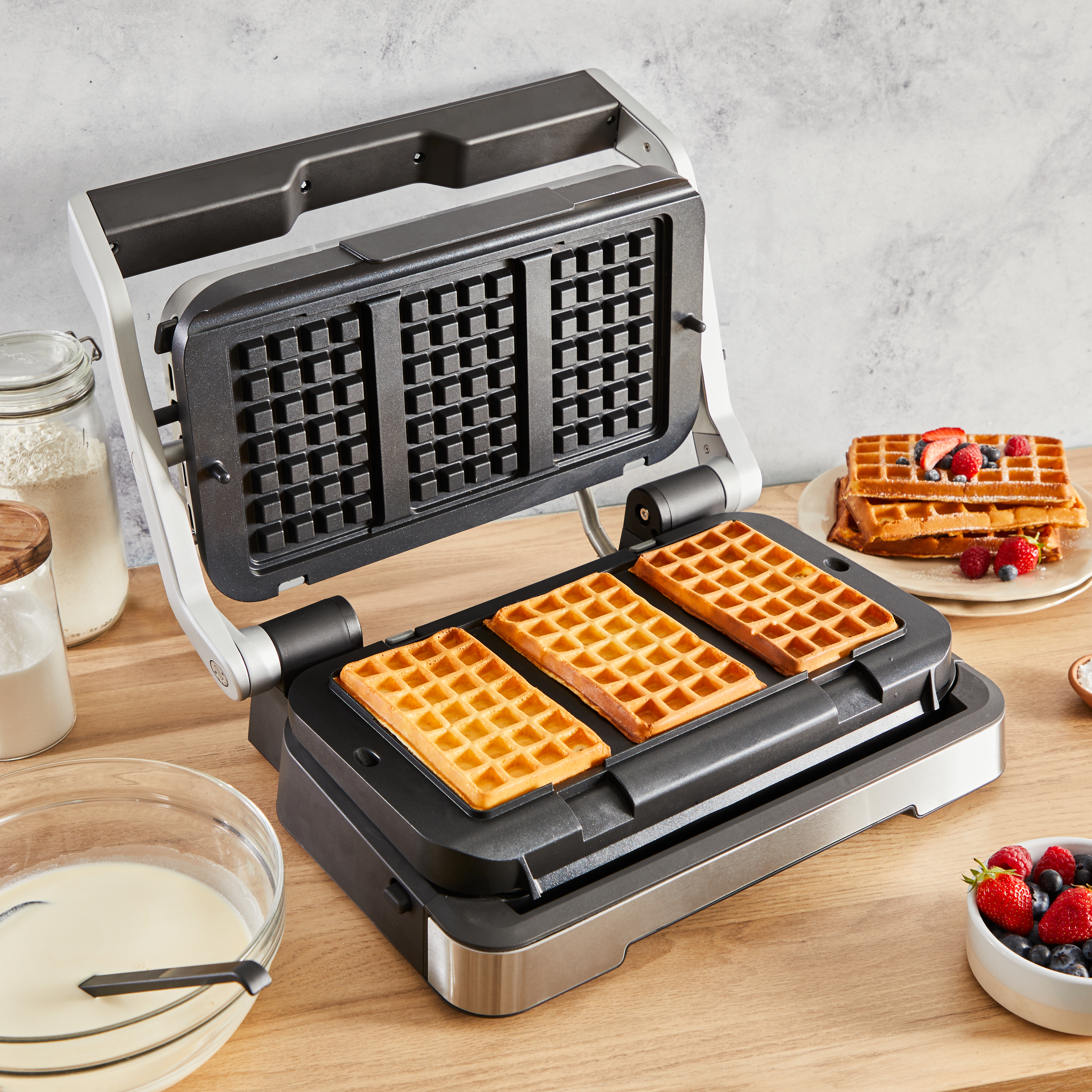 Ein Waffeleisen mit drei Waffeln darin. Auf dem Tisch befinden sich Waffeln und Zutaten.