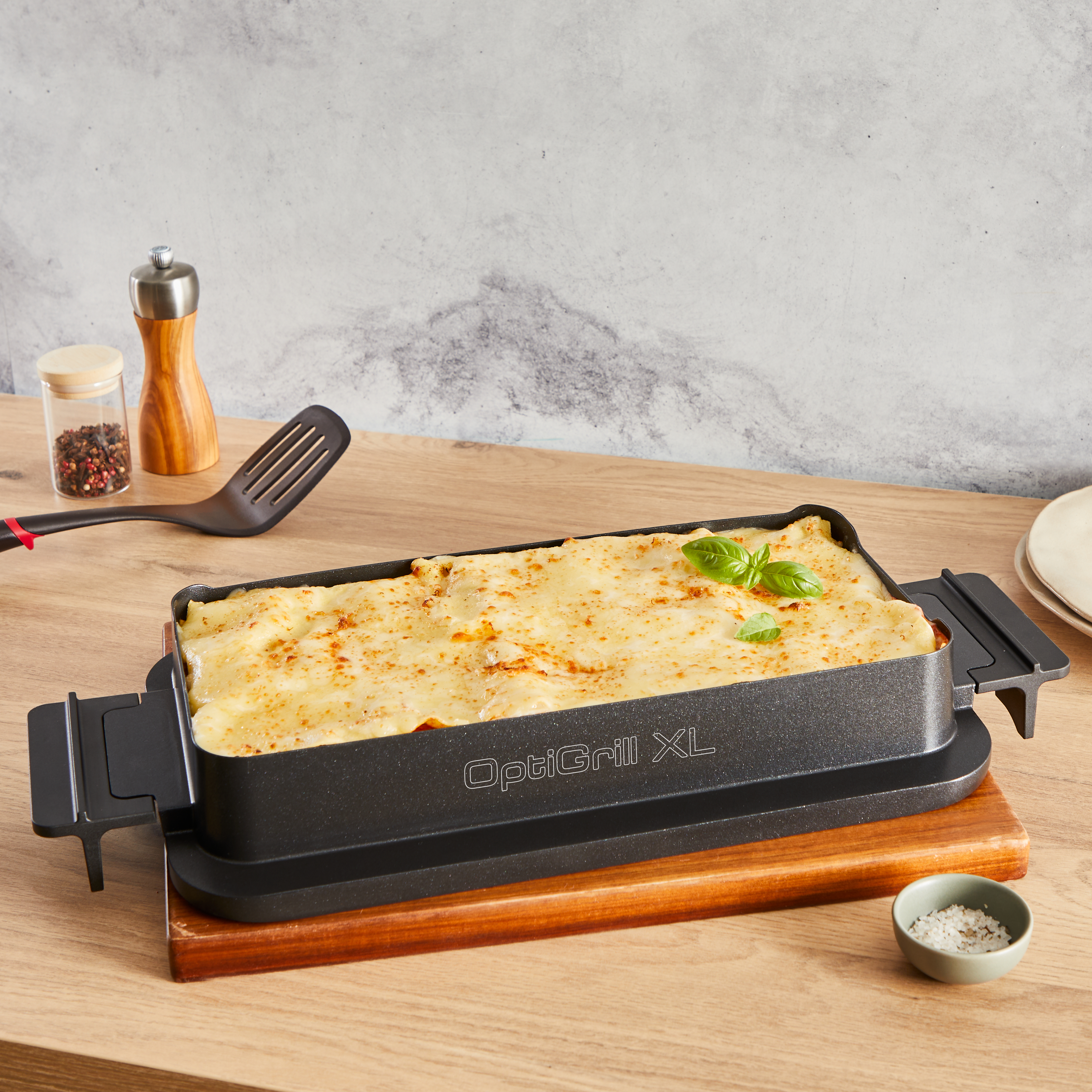 Rechteckiger OptiGrill XL mit Lasagne. Ein Holzbrett und ein Spatel liegen daneben.