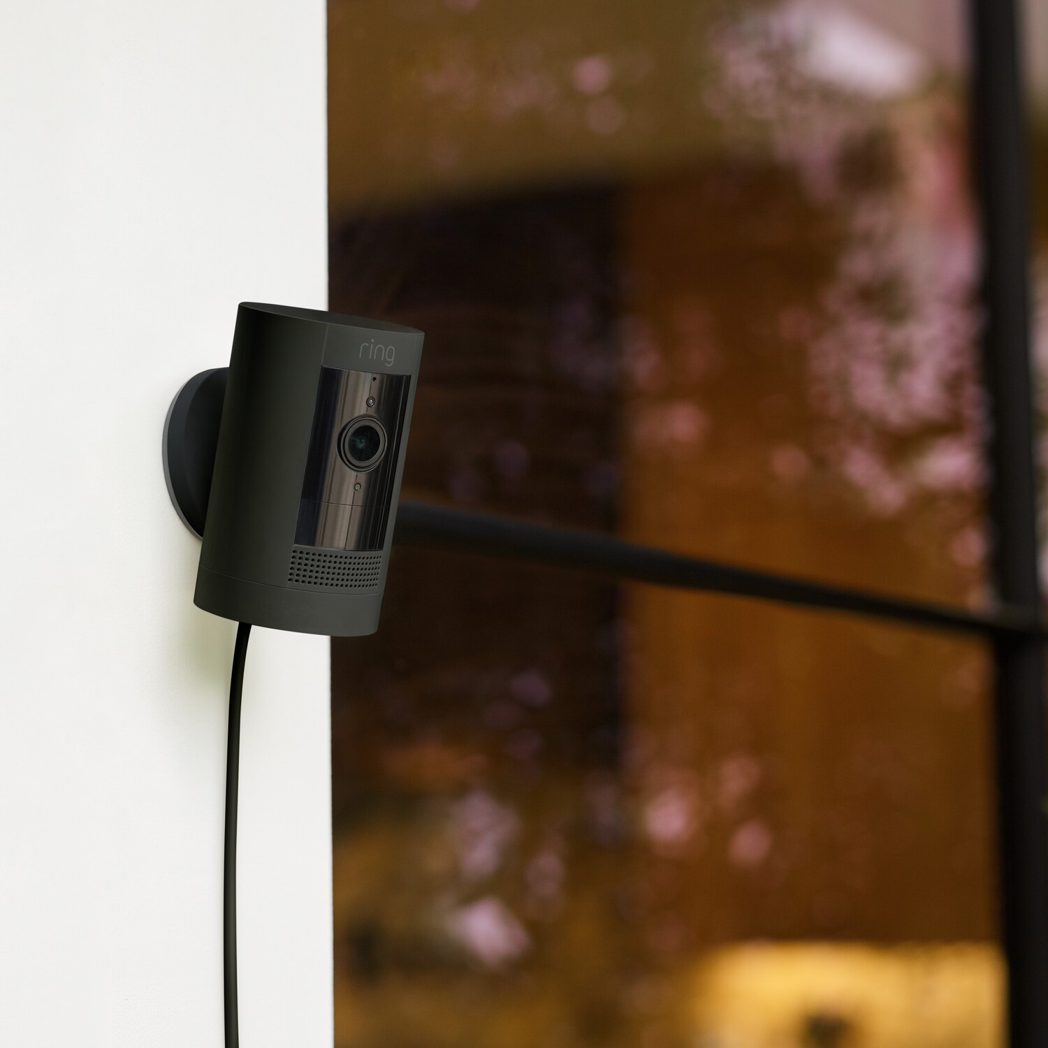 A black security camera is mounted on a white wall, with a blurred window background.
