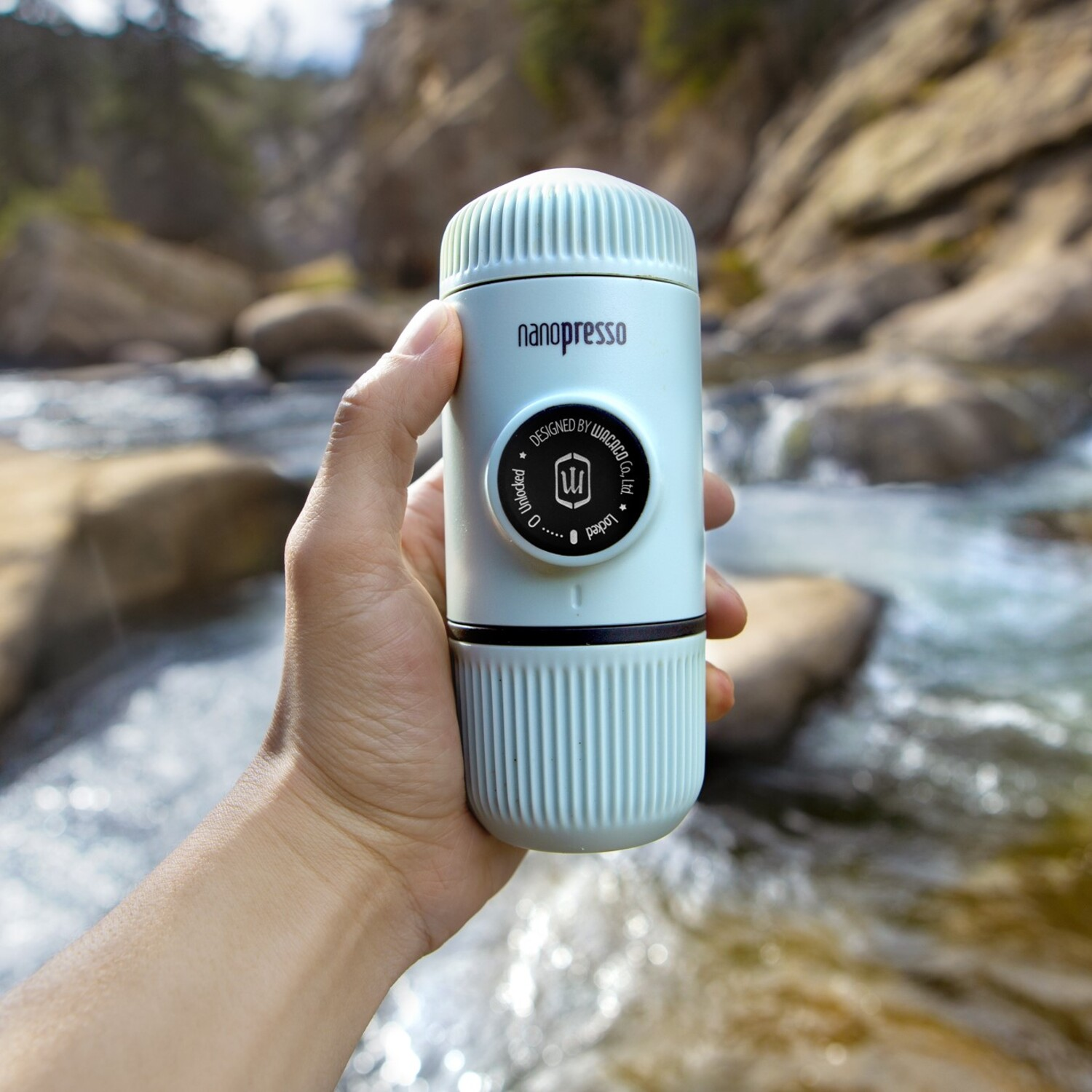 Hand die een lichtblauwe Nanopresso vasthoudt, draagbare espressomaker, met rivier op de achtergrond.