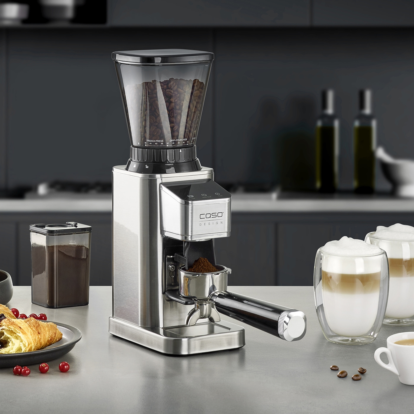 Silberne Kaffeemühle mit Bohnen, Kaffeepulver, Croissants und zwei Lattes auf grauer Arbeitsplatte.