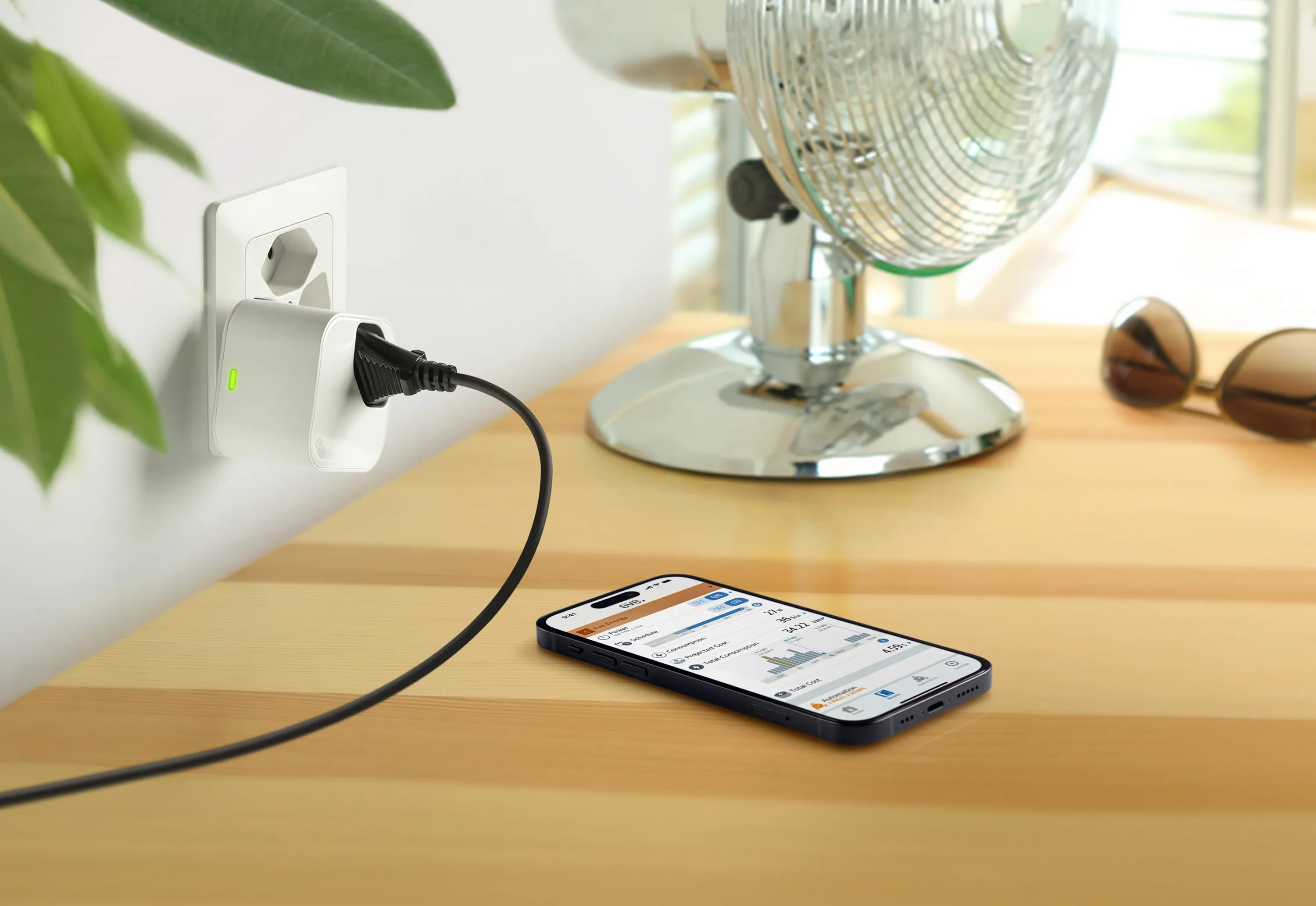 Ein Telefon lädt auf einem Holztisch. Ein Stecker steckt in einer Steckdose. Ein Ventilator und eine Sonnenbrille im Hintergrund.