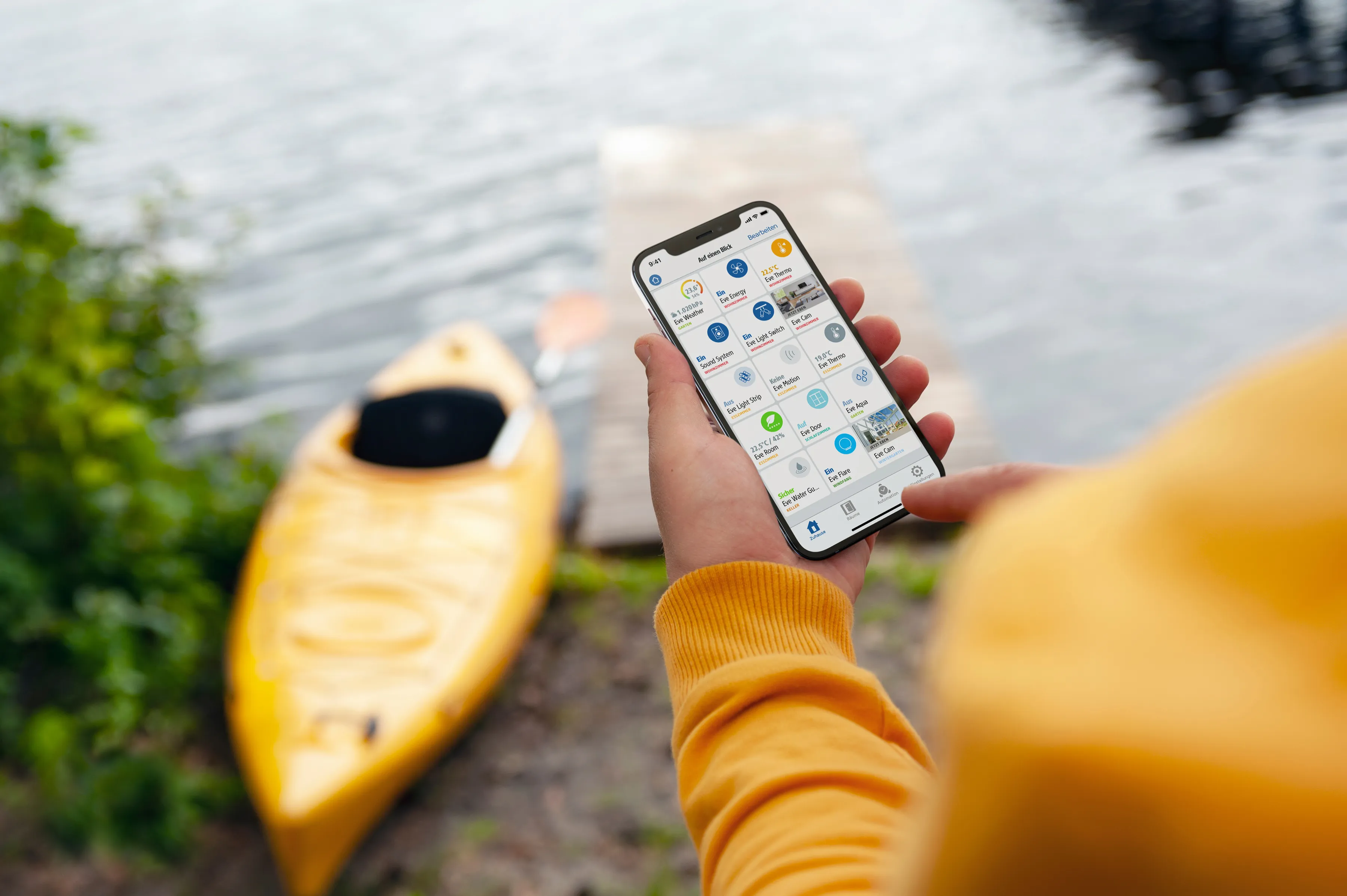 Eine Person hält ein Telefon mit einer Smart-Home-App. Ein gelbes Kajak liegt am Wasser, ein Steg im Hintergrund.
