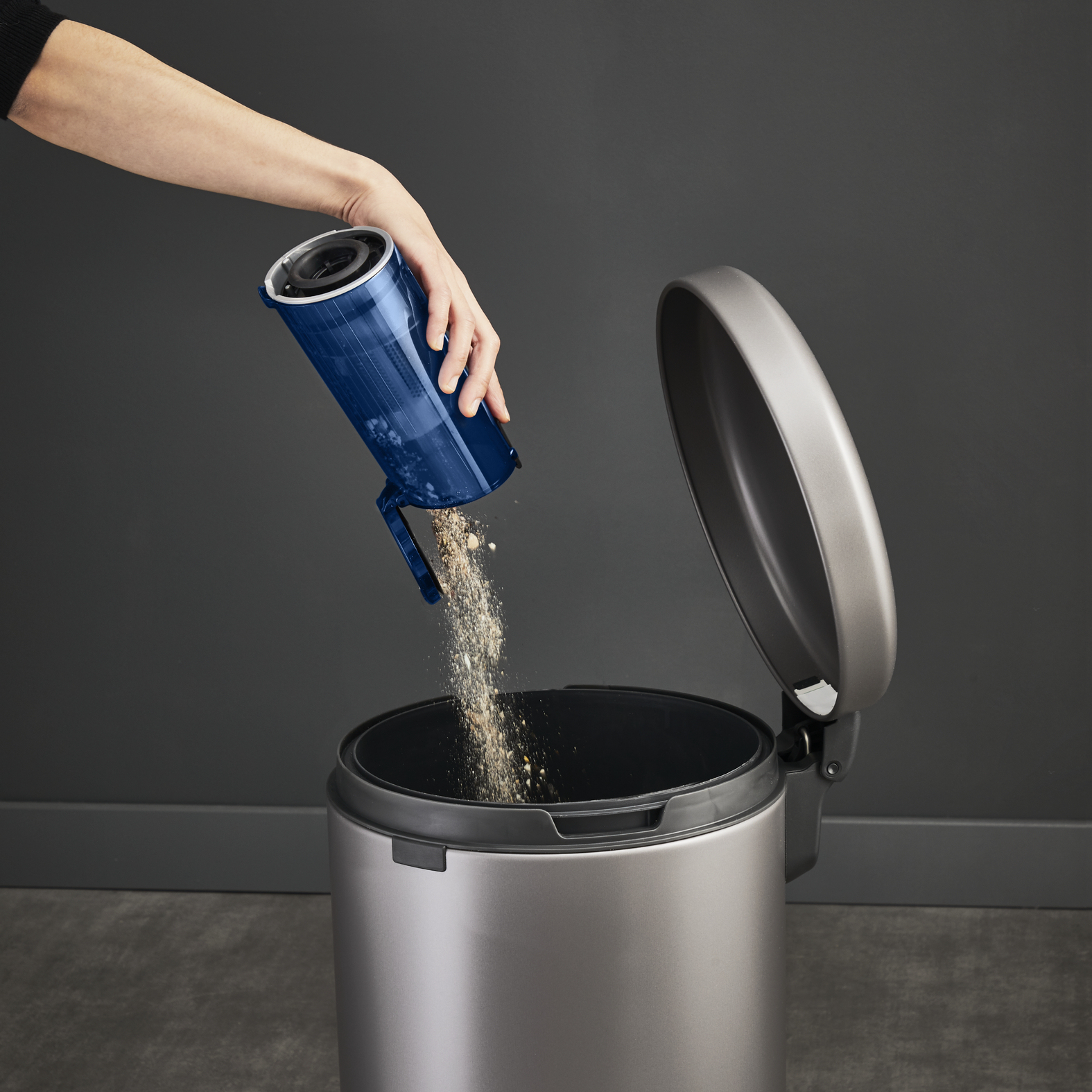 Una mano vacía un cubo de basura azul de una aspiradora en un bote de basura de acero inoxidable con la tapa abierta.