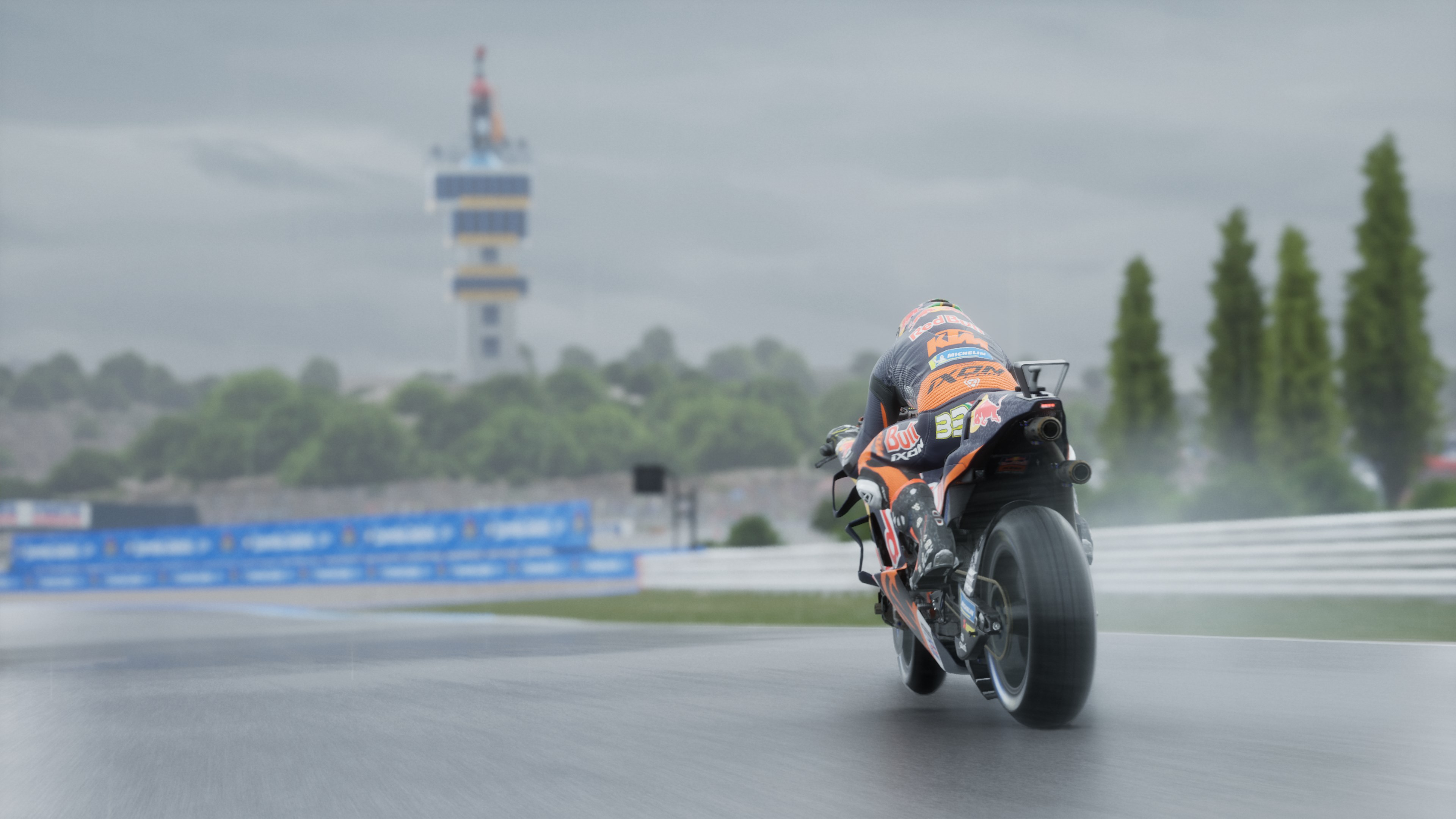 Un motocycliste sur un circuit sous la pluie, vu de dos. Une tour est en arrière-plan.