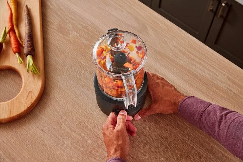 Person fügt gehacktes Gemüse einem schwarzen Food Processor auf einem Holztisch hinzu, Karotten im Hintergrund.