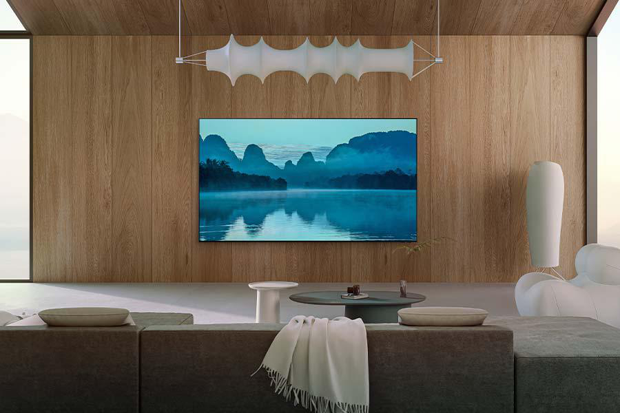 Ein Wohnzimmer mit großem Fernseher, der eine malerische Aussicht auf Berge und Wasser zeigt. Helle Holzverkleidung.