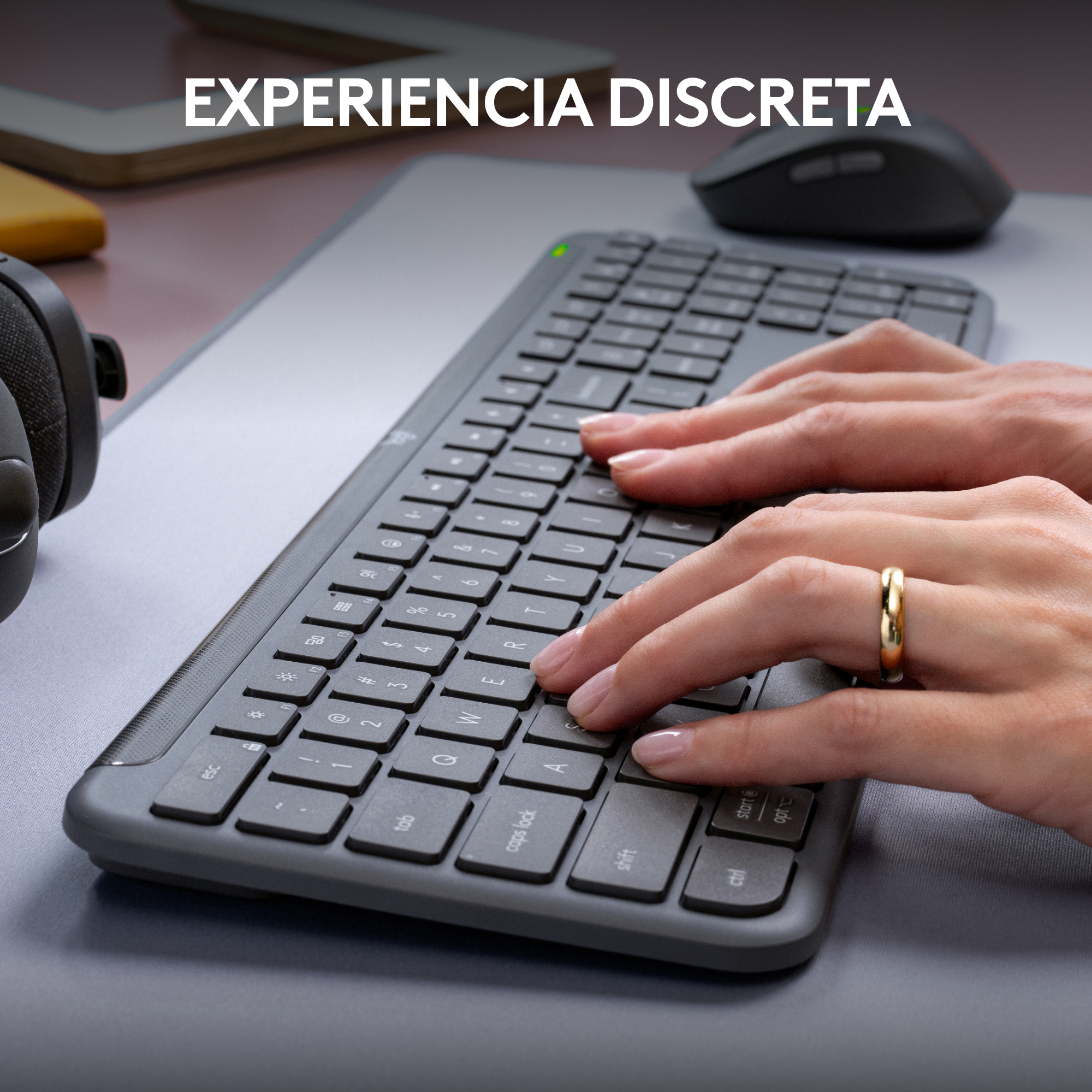 Manos tecleando en un teclado gris junto a un ratón y auriculares en un escritorio.