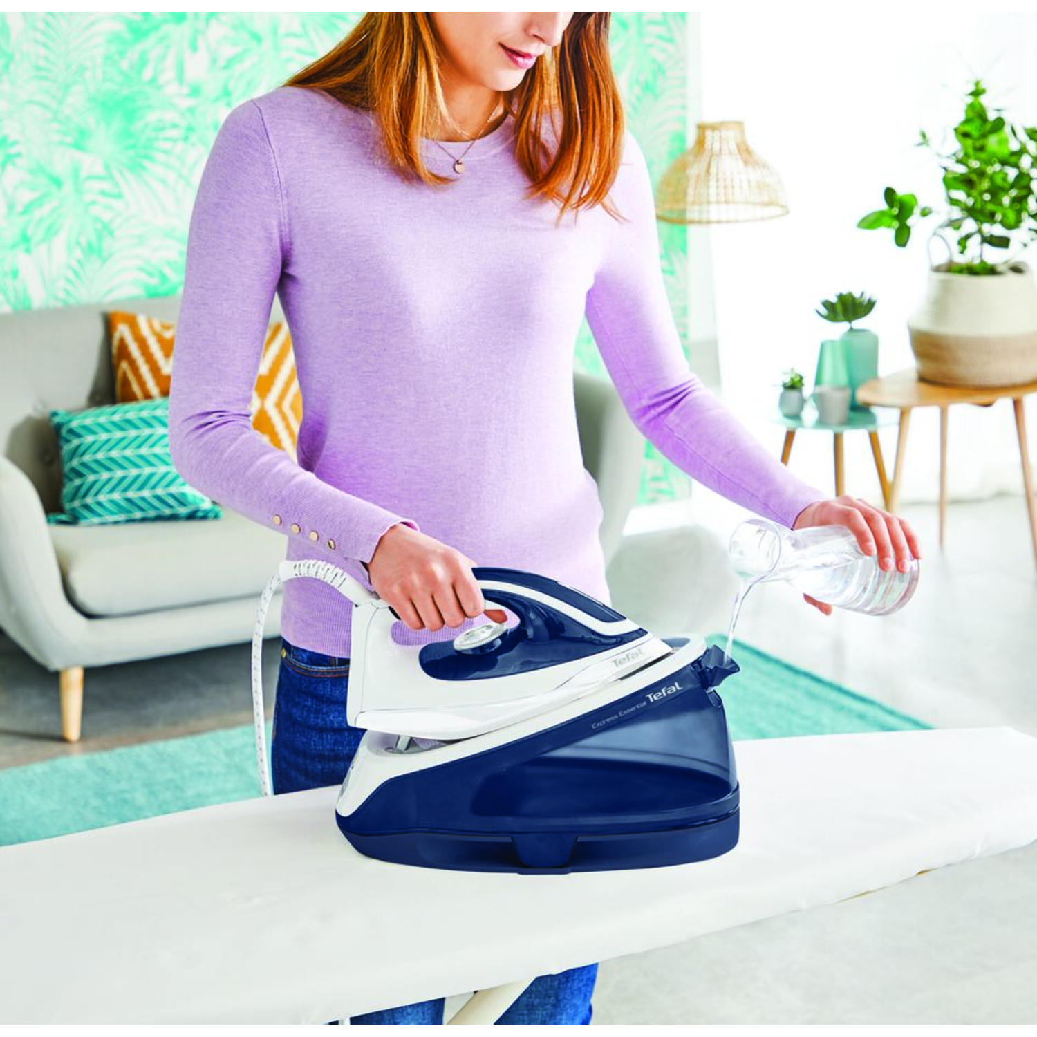 A woman pouring water into a white and blue iron, set on an ironing board, in a living room.