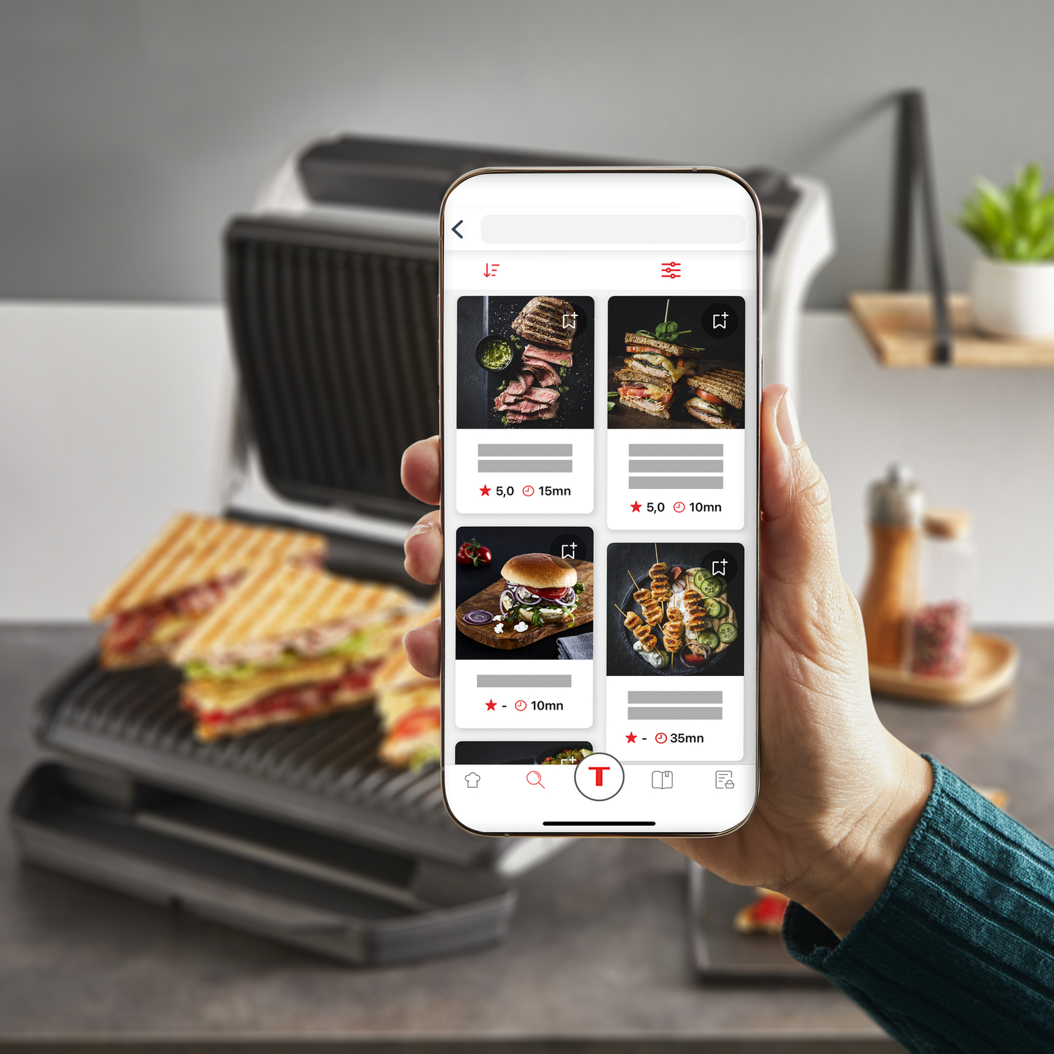A person holding a phone displaying a food recipe app. A grill and cooked food are in the background.