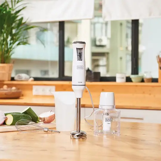 Biały blender ręczny i akcesoria na drewnianym blacie z warzywami.