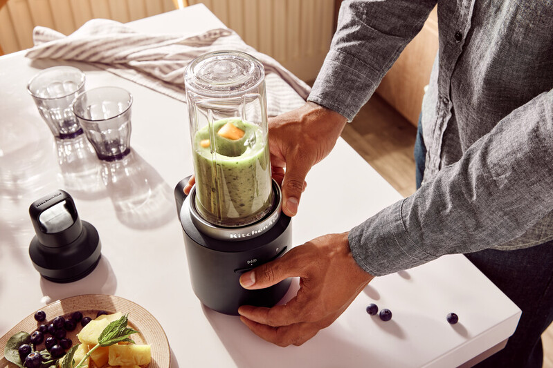 Person montiert einen schwarzen und silbernen KitchenAid-Mixer. Zutaten und Gläser liegen auf dem Tisch.