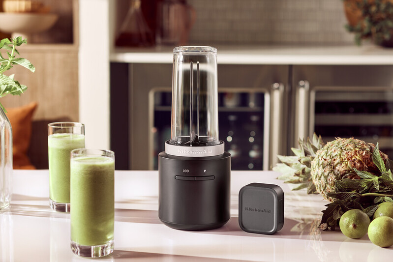 Ein schwarzer Mixer mit zwei grünen Smoothies und Früchten.