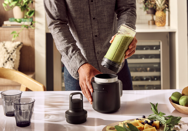 Eine Person montiert einen schwarzen Mixer mit einem grünen Smoothie auf einem weißen Tisch.