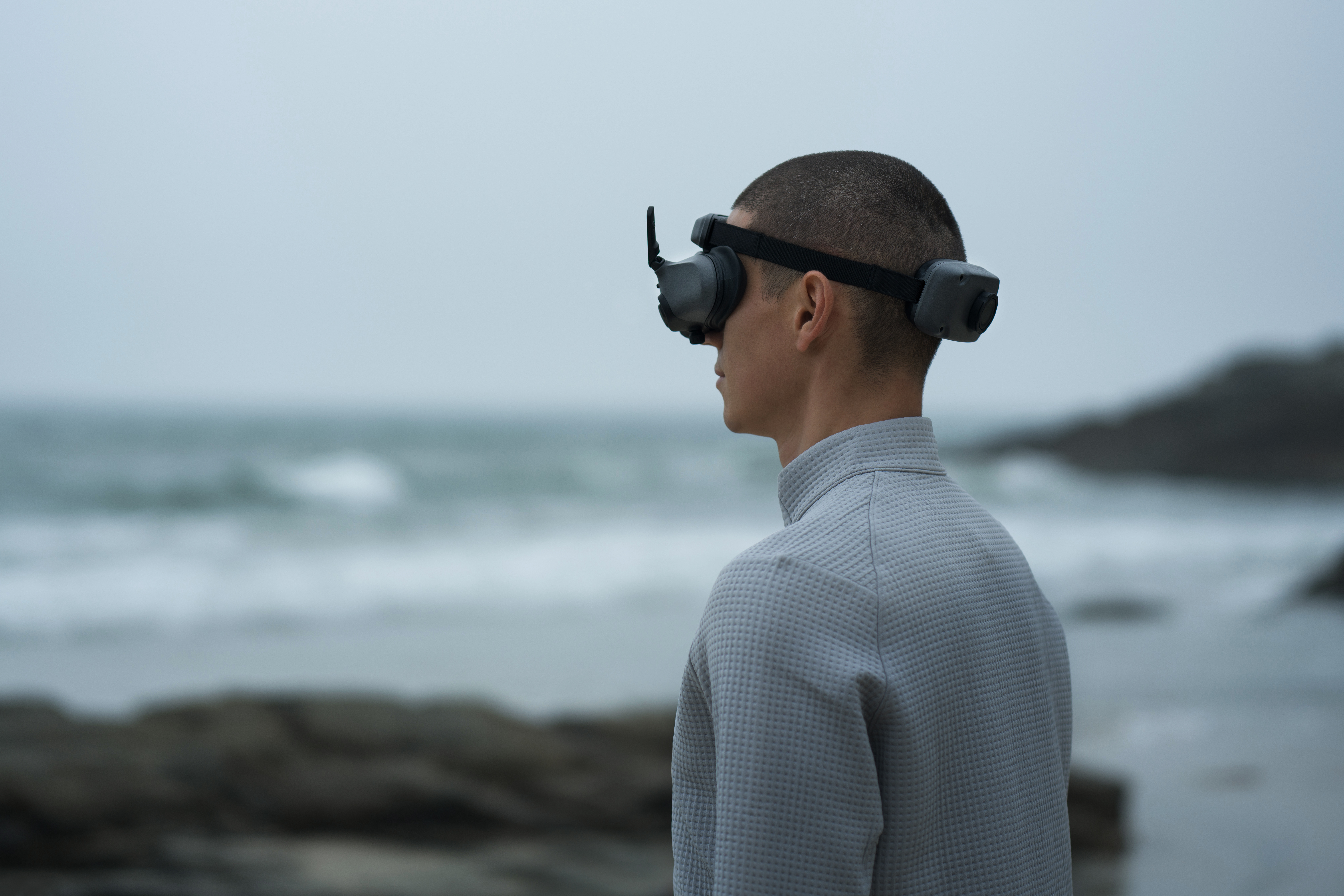 Homme avec casque VR sur une plage, regardant la mer, ciel nuageux. Vêtements gris.