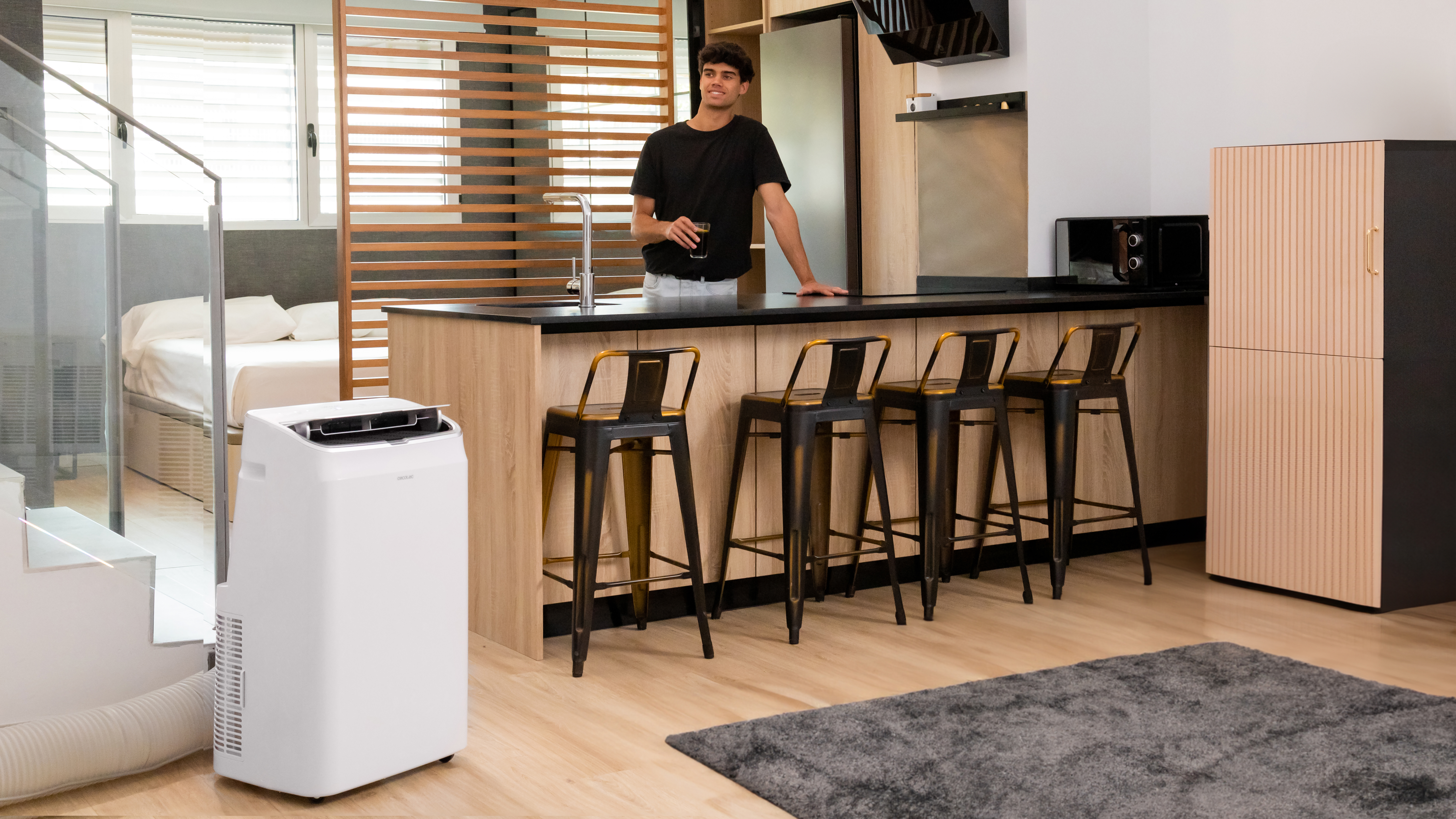 Interior de apartamento moderno con un hombre sosteniendo una bebida. Unidad de AC, cocina y sala de estar visibles.