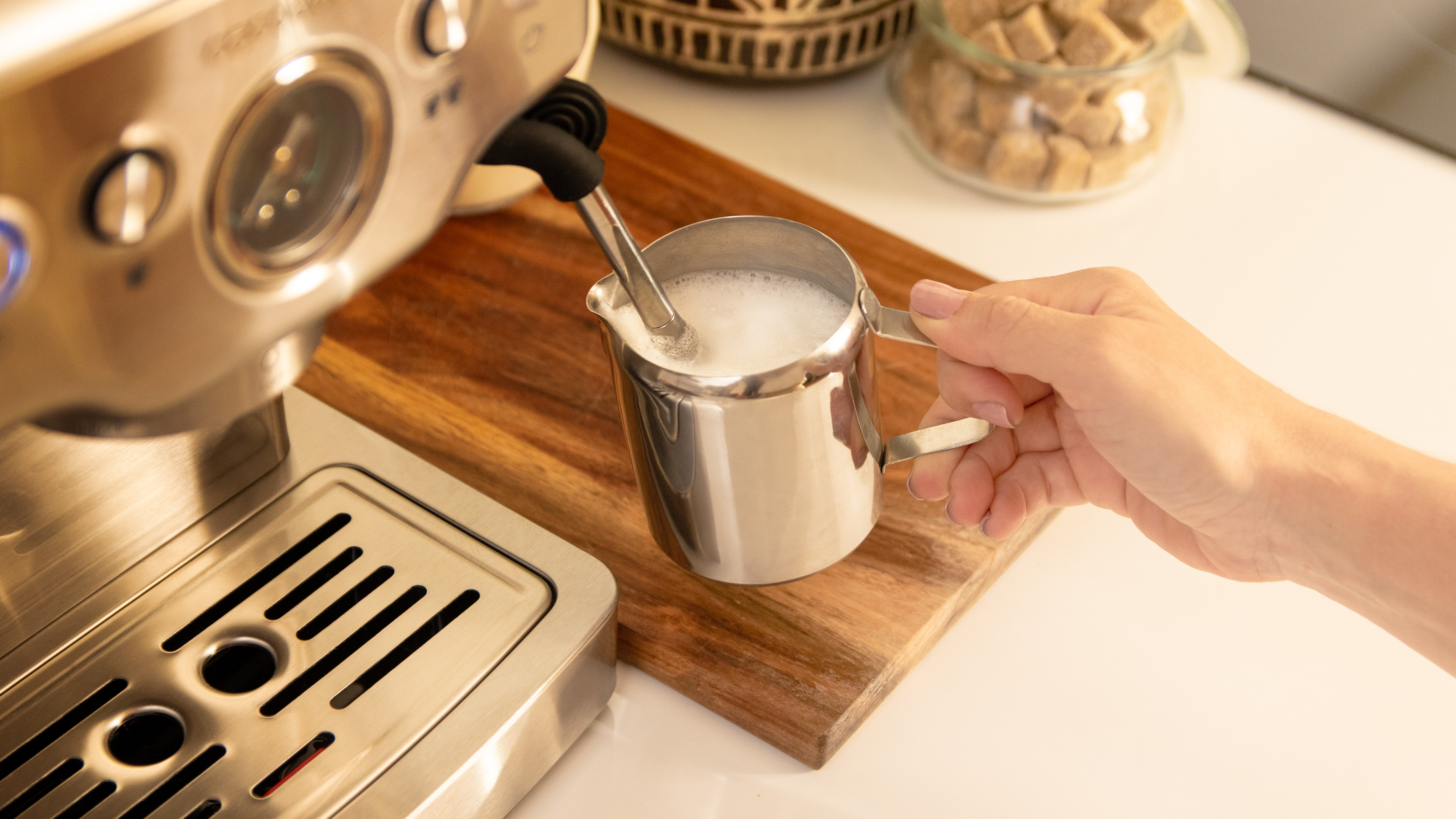 La mano de una persona sosteniendo una jarra de acero inoxidable para espumar leche junto a una máquina de espresso.