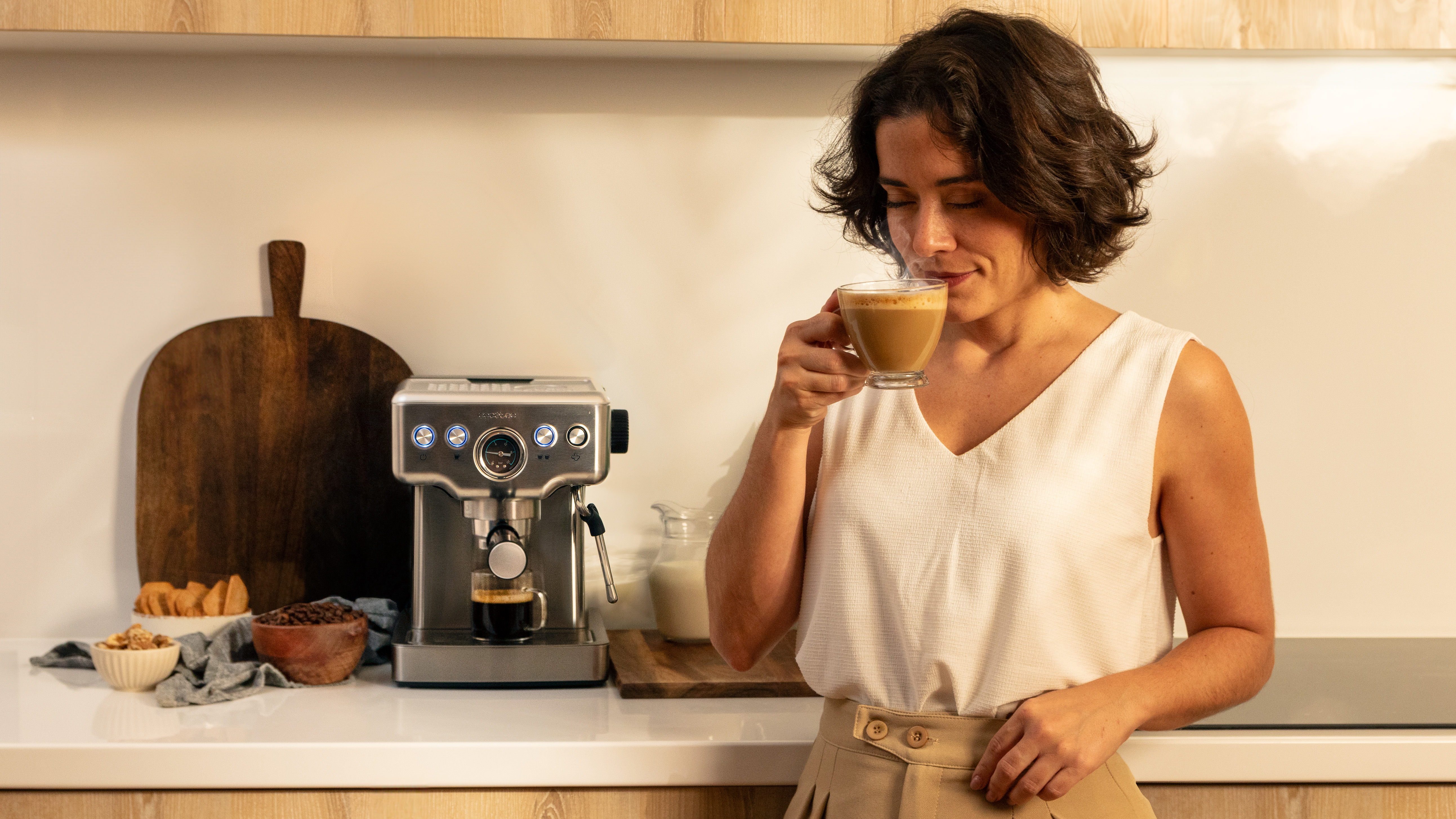Una mujer bebe café de una taza de vidrio. Cafetera, tabla y cuenco de nueces cerca.