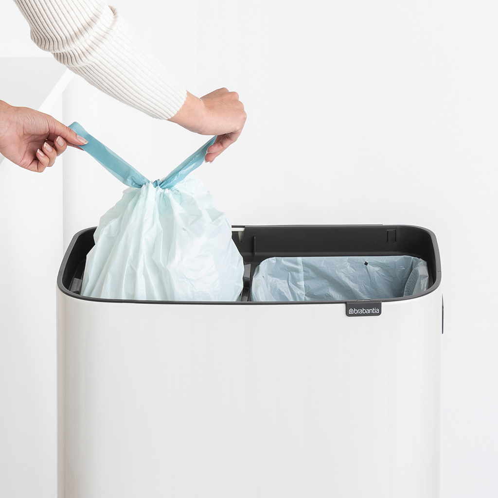 Hände legen einen hellblauen Müllsack in einen weiß-schwarzen Mülleimer. Ein weiterer Sack drin.