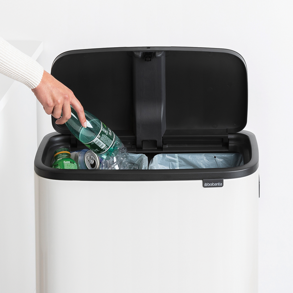 Die Hand einer Frau, die eine grüne Plastikflasche in einen weißen Mülleimer mit zwei Fächern legt.