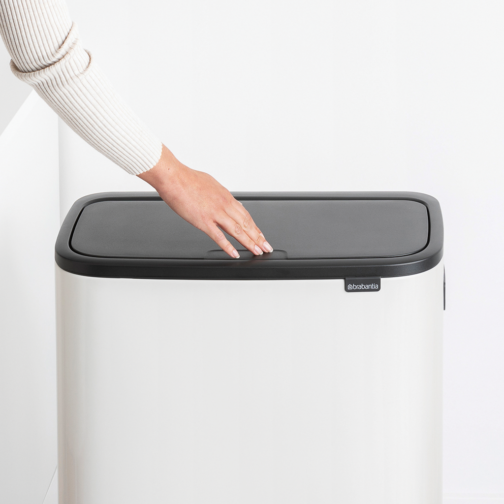 Eine Hand berührt den Deckel eines weißen Brabantia-Mülleimers, ein schwarzer Deckel.