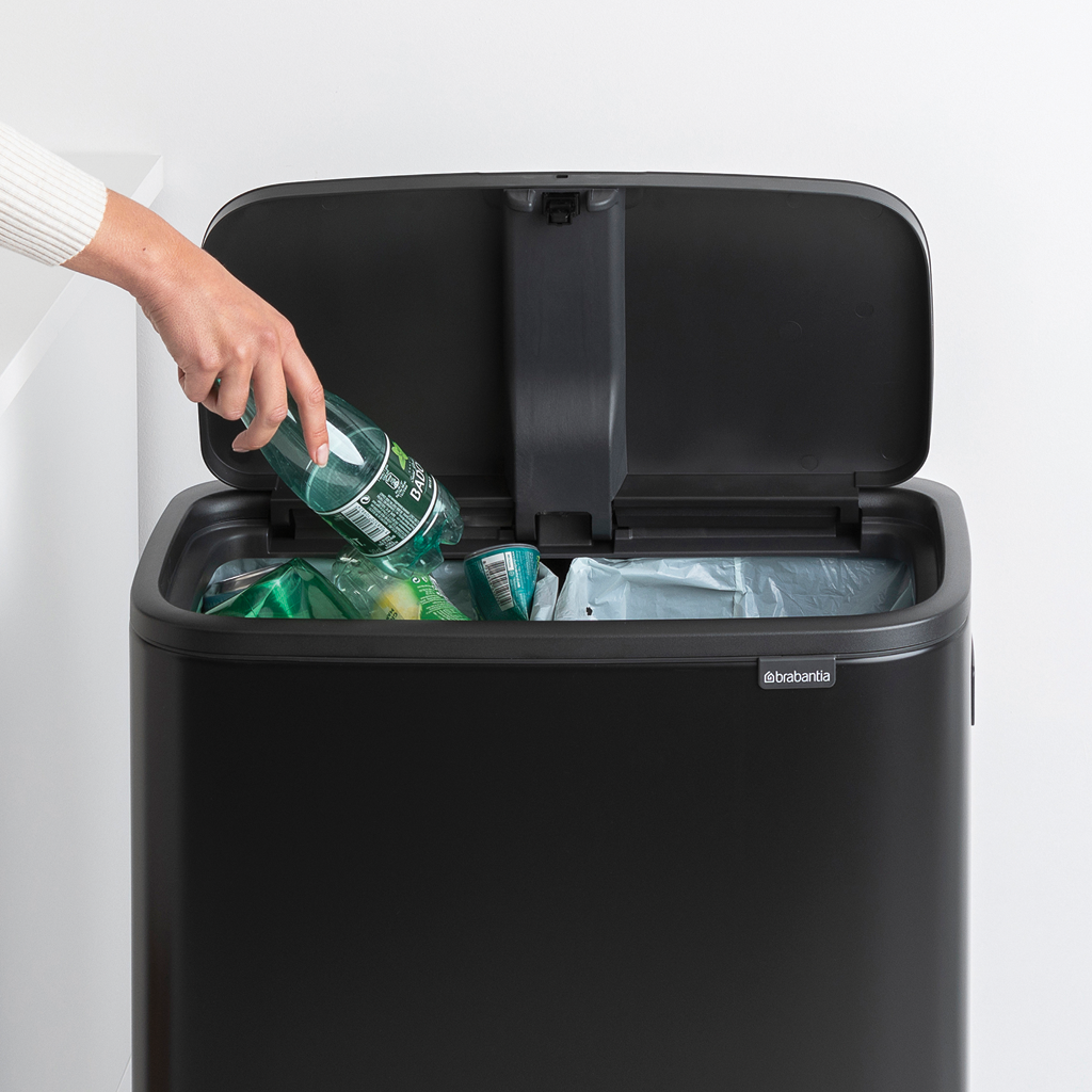 Eine Person wirft eine Plastikflasche in einen schwarzen Mülleimer. Der Deckel ist geöffnet und zeigt Müllsäcke.