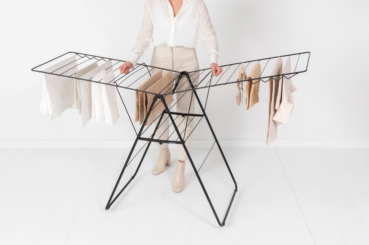 Eine Frau steht neben einem schwarzen Wäscheständer mit Kleidung. Weißer Wandhintergrund.