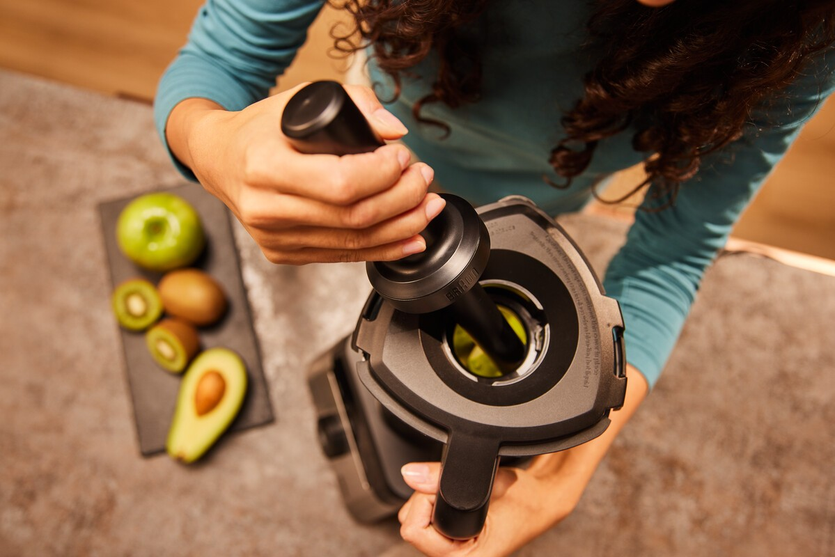 Eine Person benutzt einen Entsafter mit Früchten wie Kiwi, Apfel und Avocado daneben.