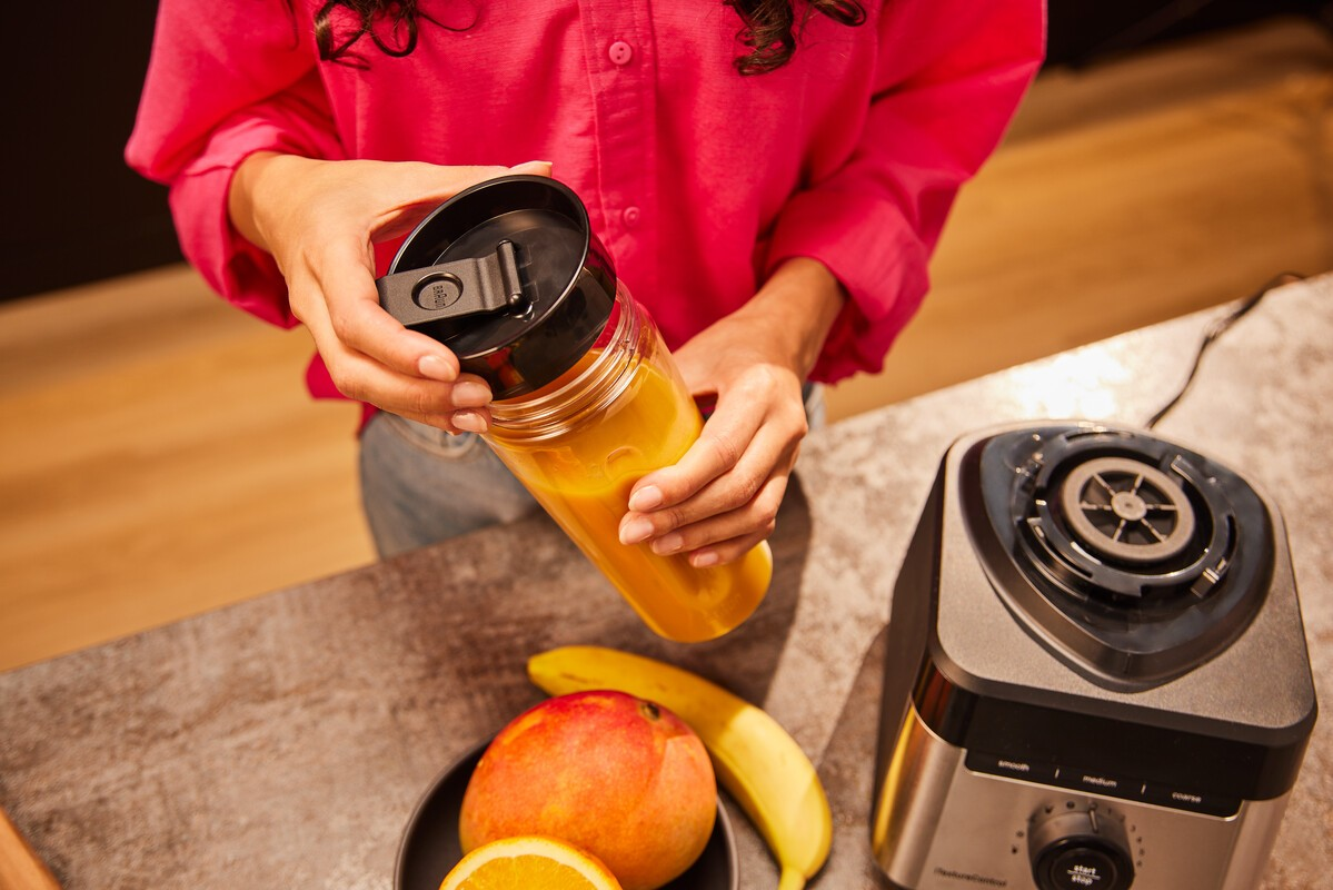 Eine Person hält eine Mixerflasche mit orangem Inhalt neben einem Mixer und Obst.