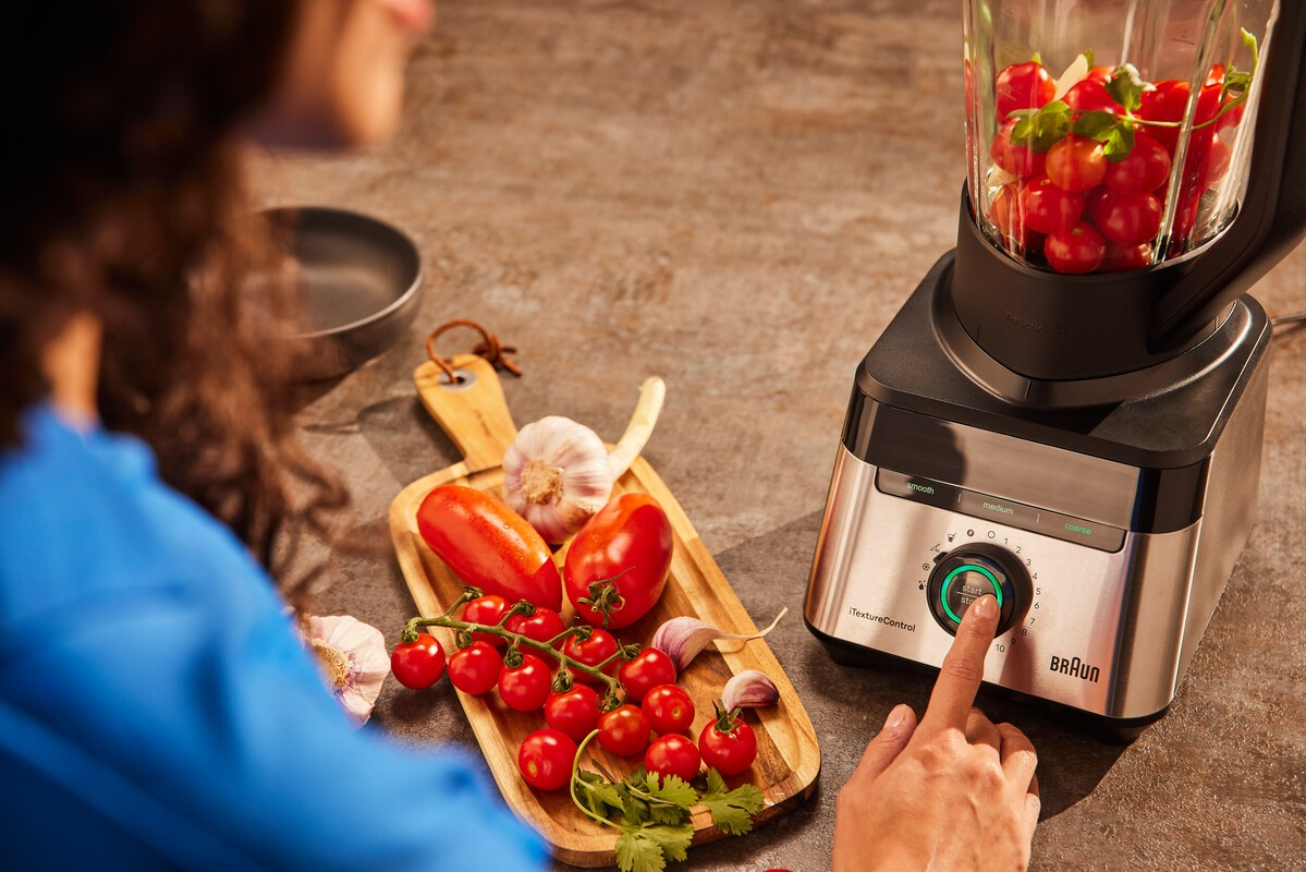 Die Hand einer Person dreht den Drehregler eines Mixers mit Tomaten und Kräutern.