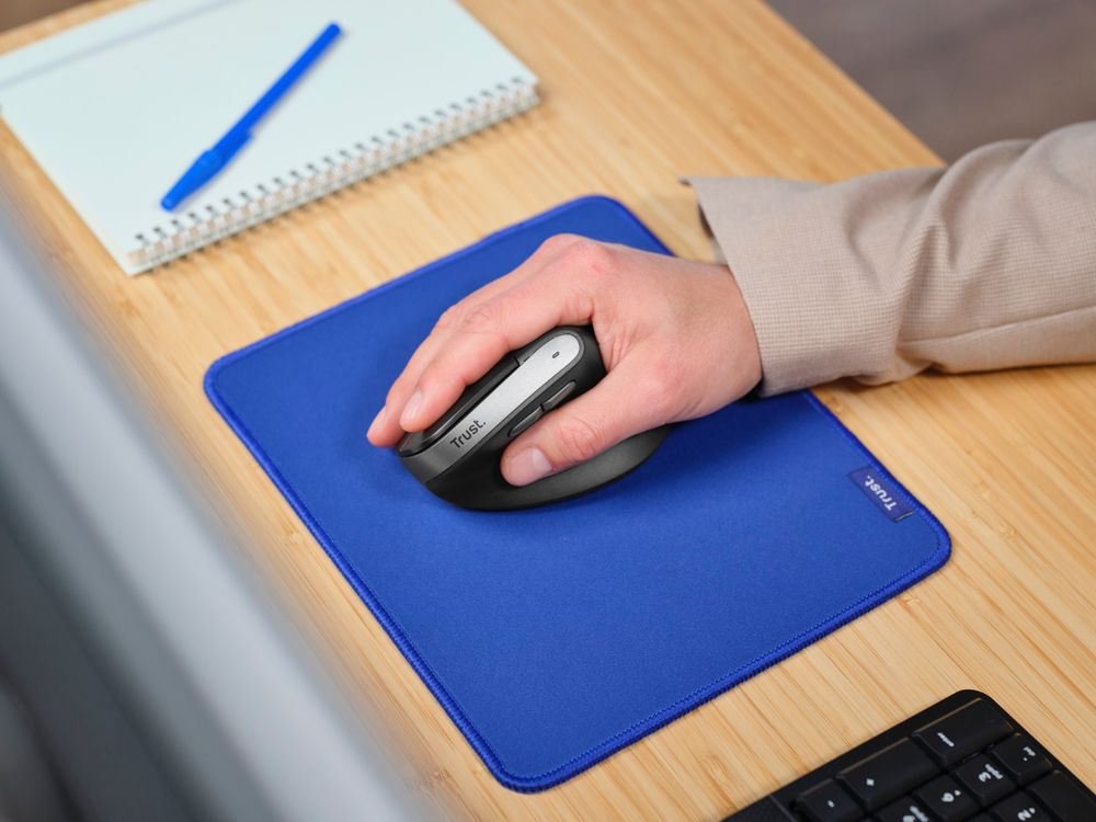 Mano usando un ratón negro y plateado en una alfombrilla azul, con un cuaderno y bolígrafo.