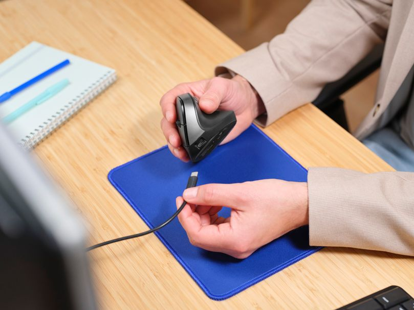 Una persona sosteniendo un ratón y un cable USB-C, sobre un escritorio con una alfombrilla azul.