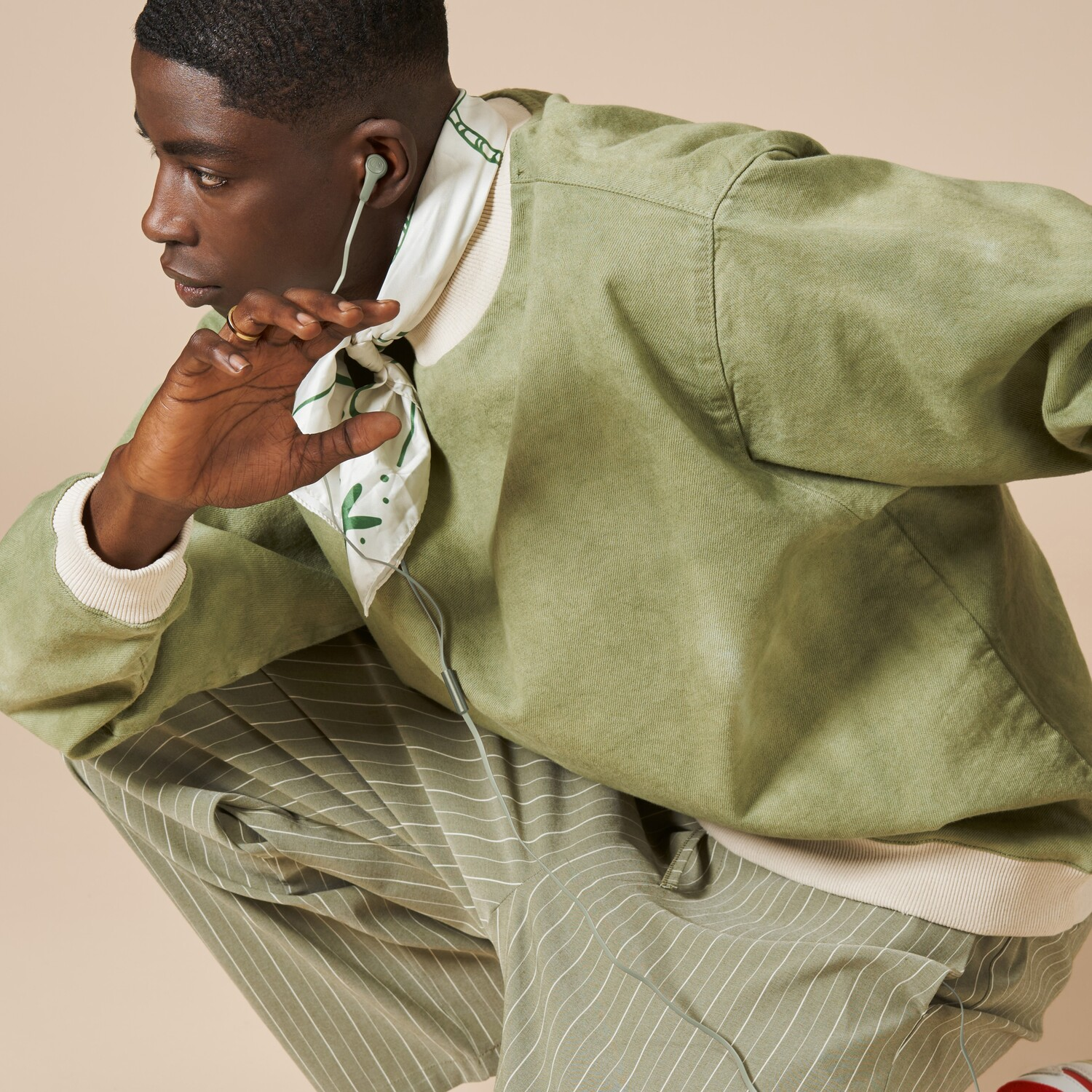 Een jonge zwarte man met oortelefoons, een groene jas en een gestreepte broek, hurkend. Beige achtergrond.