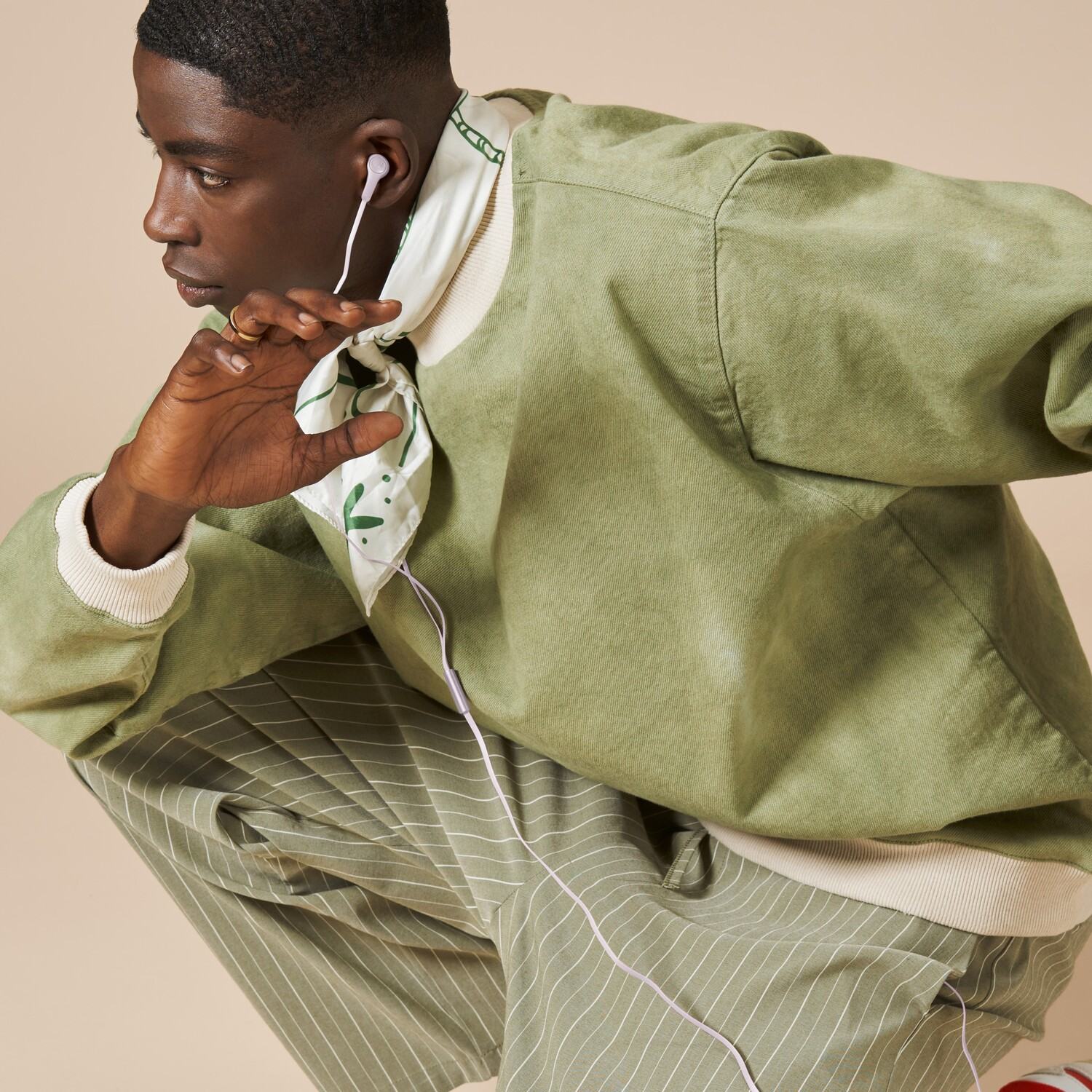 A man in green jacket and pants, wearing earbuds, is squatting.