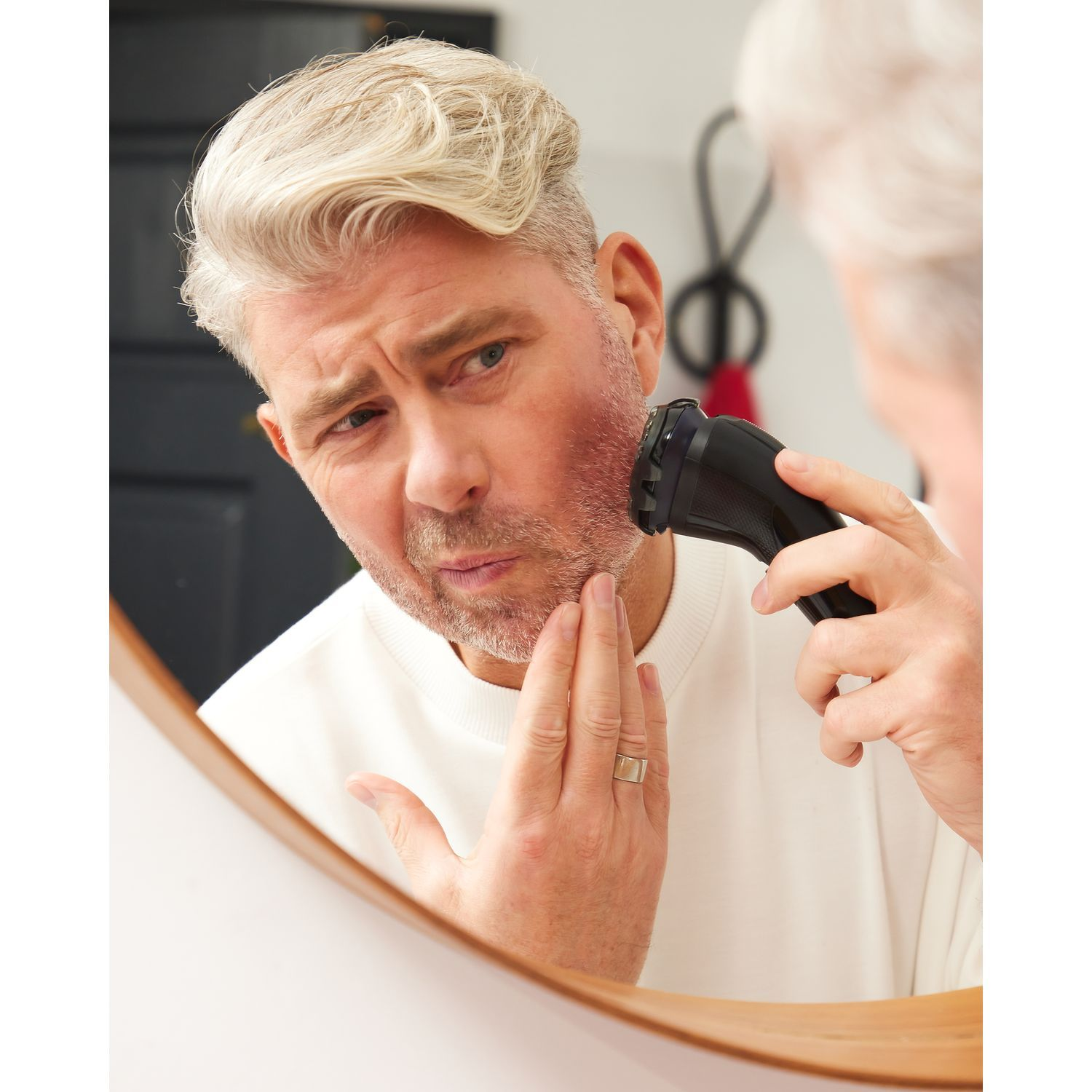 Man shaves his beard, looking in a mirror. He holds the shaver to his cheek.