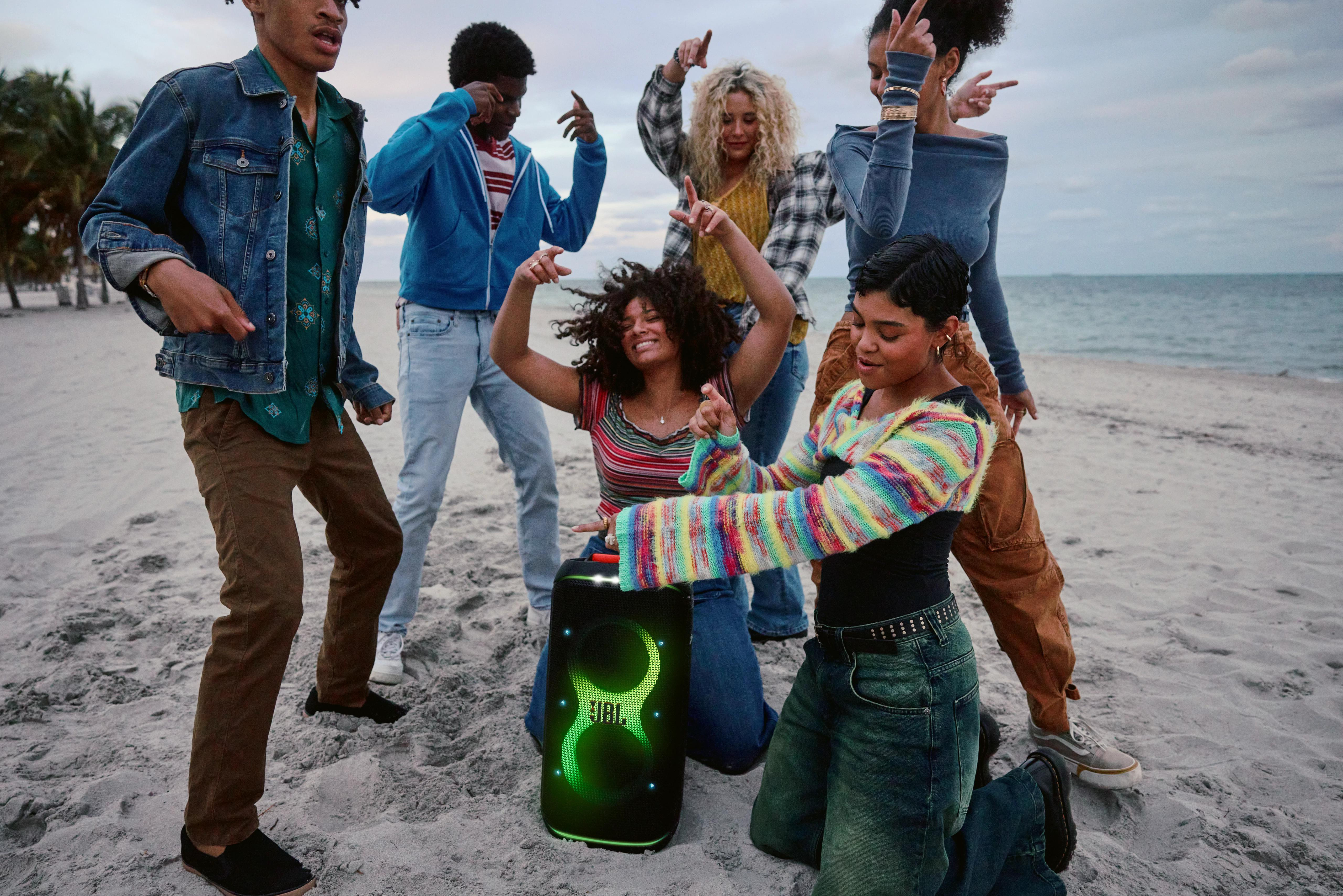 Eine Gruppe von Menschen tanzt mit einem JBL-Lautsprecher am Strand. Der Himmel ist bewölkt.